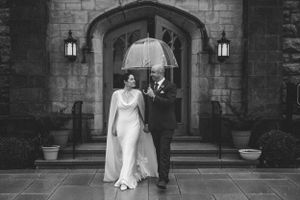 A couple stands under a stone archway sharing a white umbrella on their rainy wedding day.