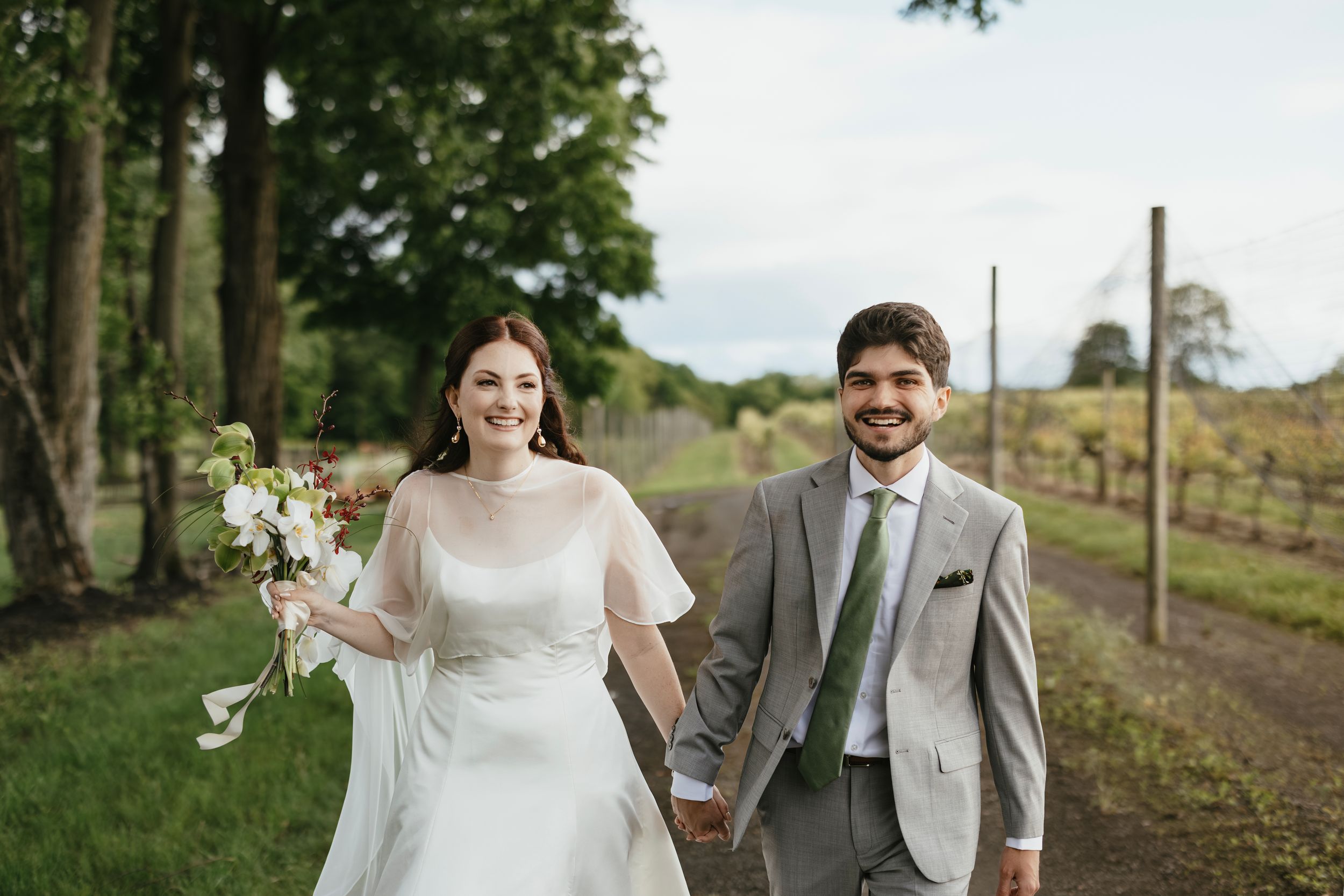 A couple walks hand in hand on a tree-lined path during their wedding celebration on a sunny day.