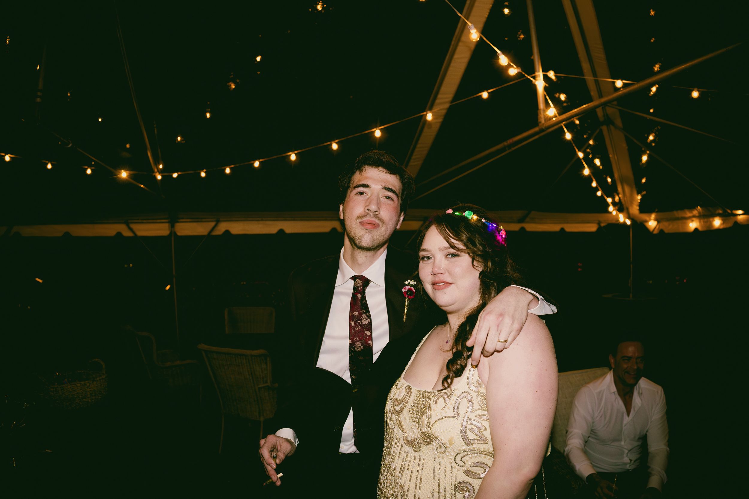 Wedding reception guests dancing and celebrating under warm string lights in a dimly lit venue at nighttime.