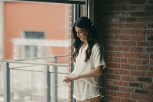 Someone in casual white attire leans against a brick wall by a window while looking down at their phone.