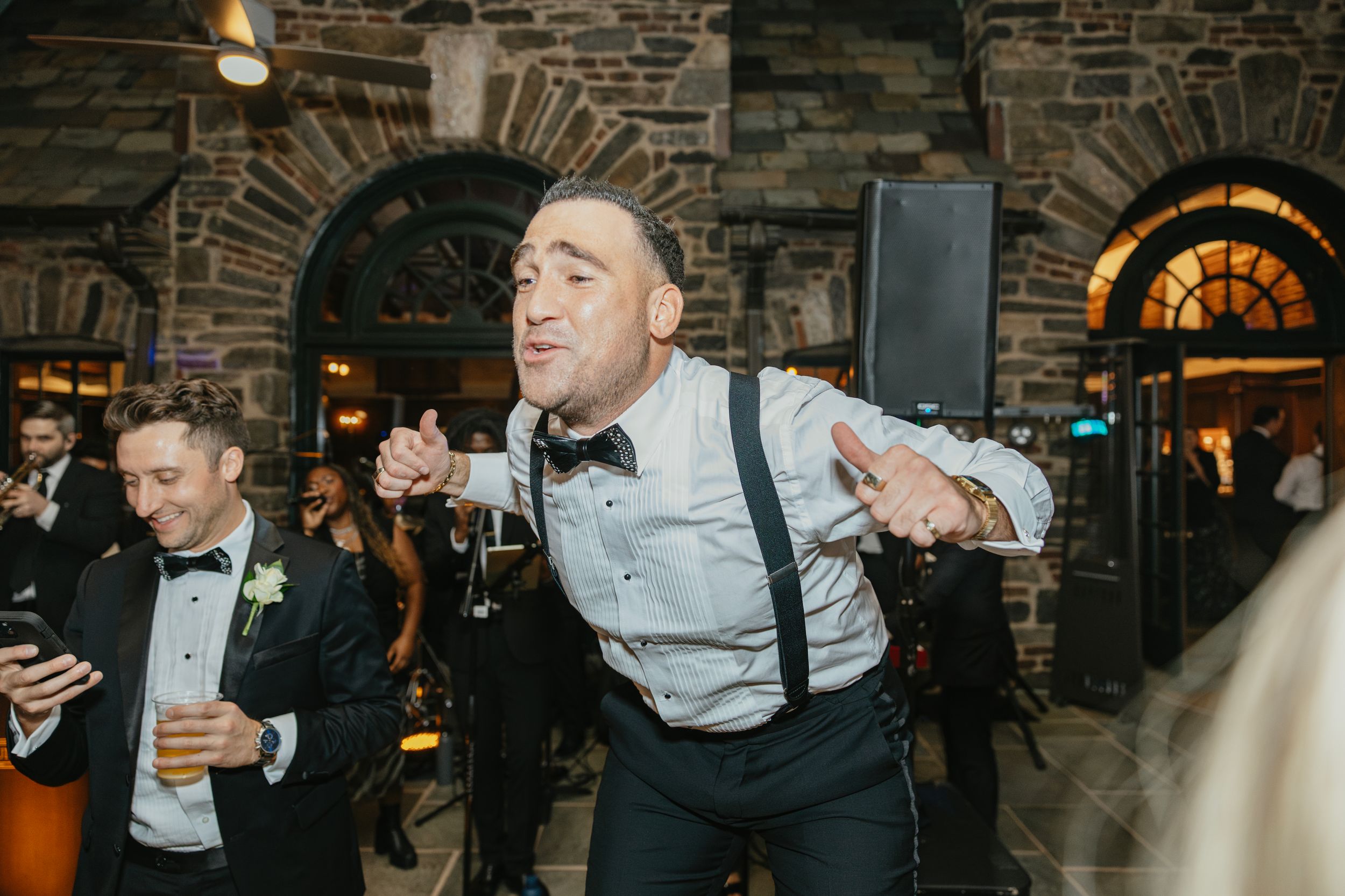 Wedding guests dancing and celebrating at a stone-walled venue with arched doorways and warm lighting.