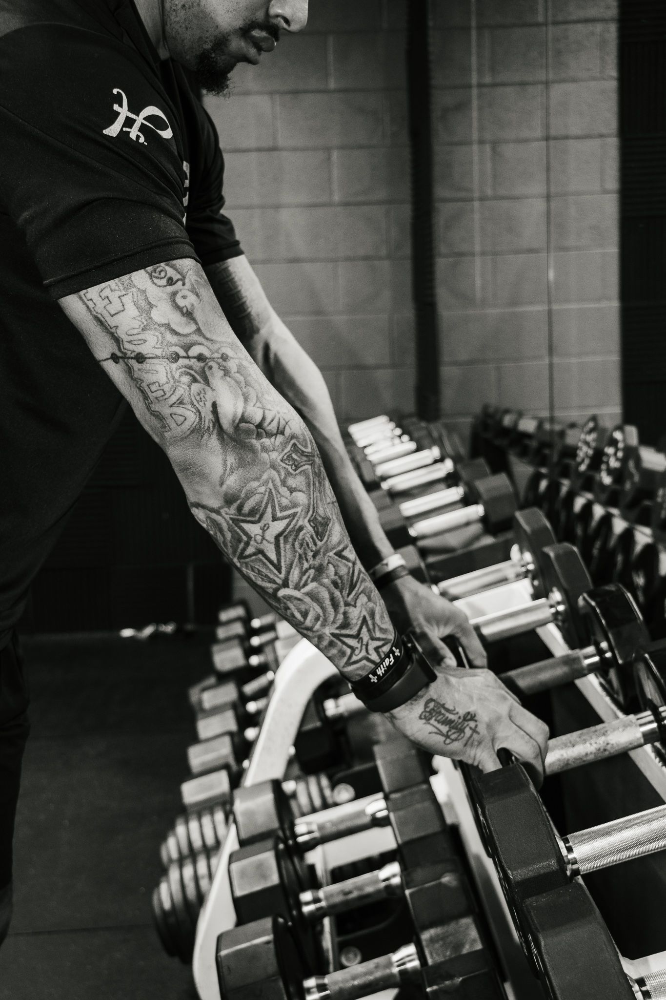 Tattooed arms reach for dumbbells in a gym, showing detailed ink and a focused stance in front of a weight rack.