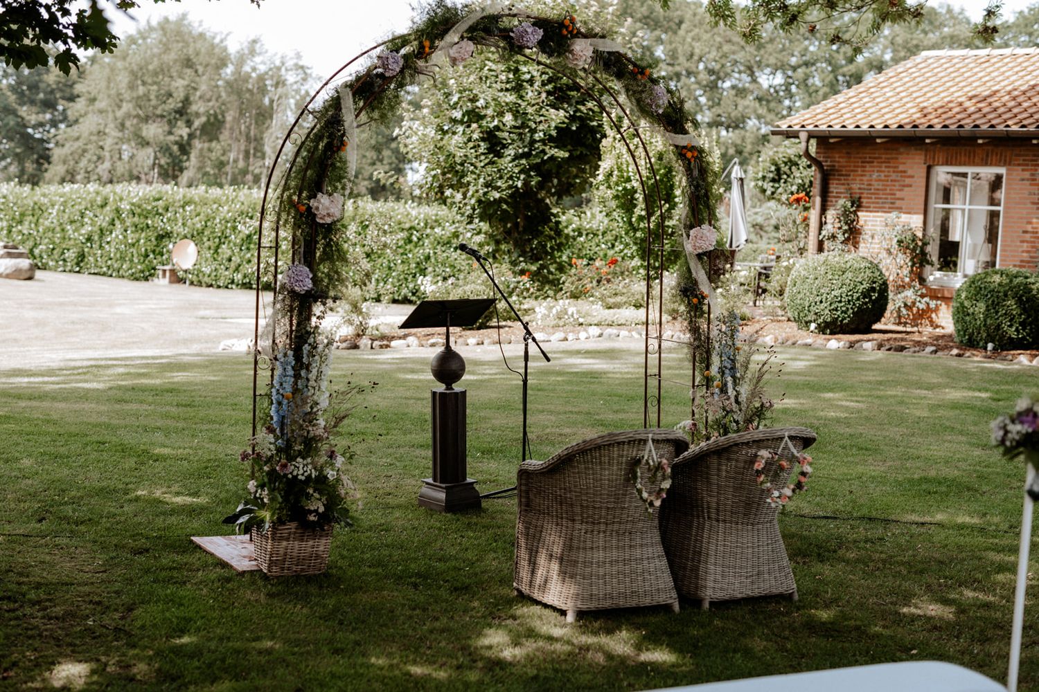 A garden wedding arch decorated with greenery and wicker chairs beneath it.