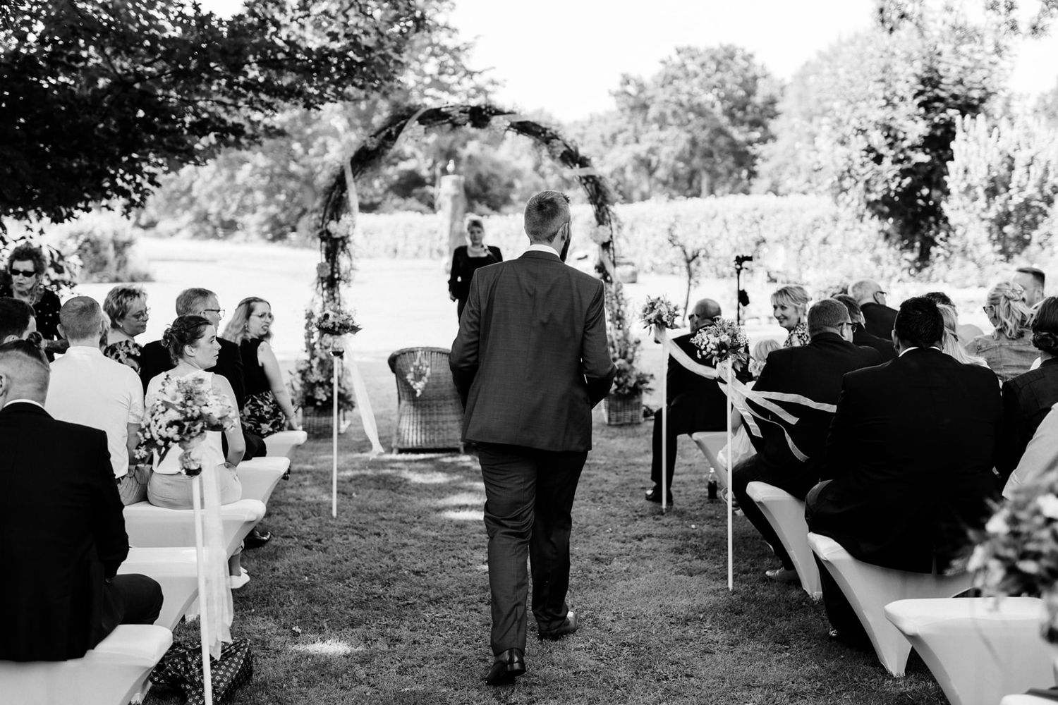 Black and white photo of outdoor wedding processional with floral arch.
