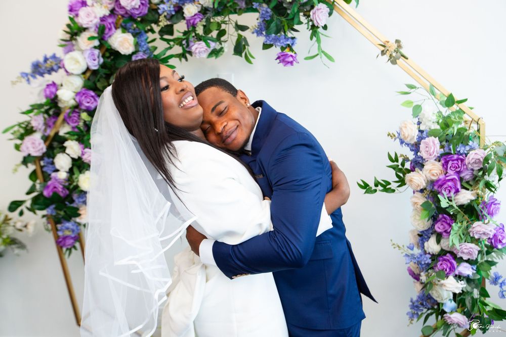 A joyful wedding couple embraces under a floral arch decorated with purple and white flowers.