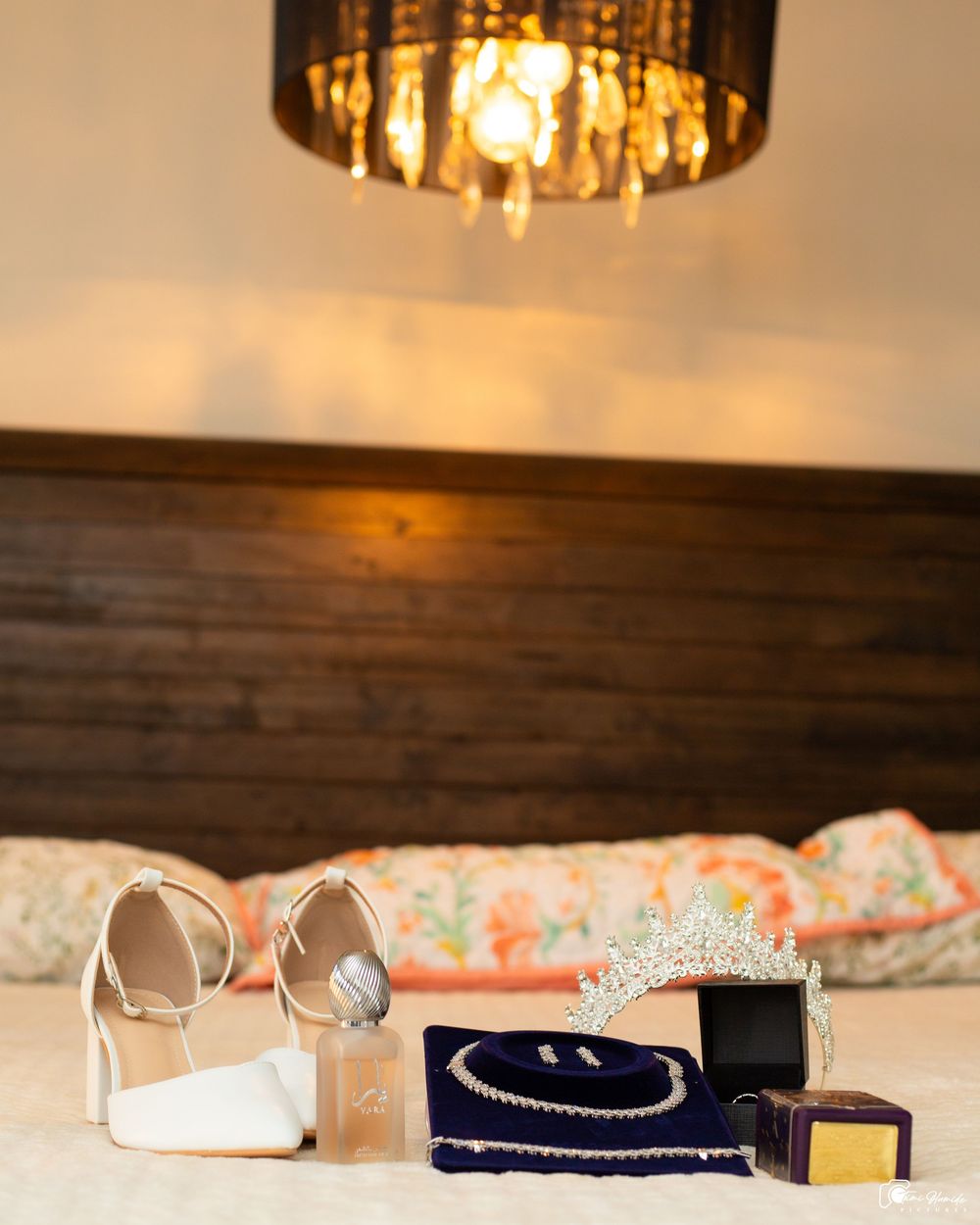 Bridal accessories display featuring white heels, tiara, pearl jewelry in velvet box, and perfume on bed under chandelier.