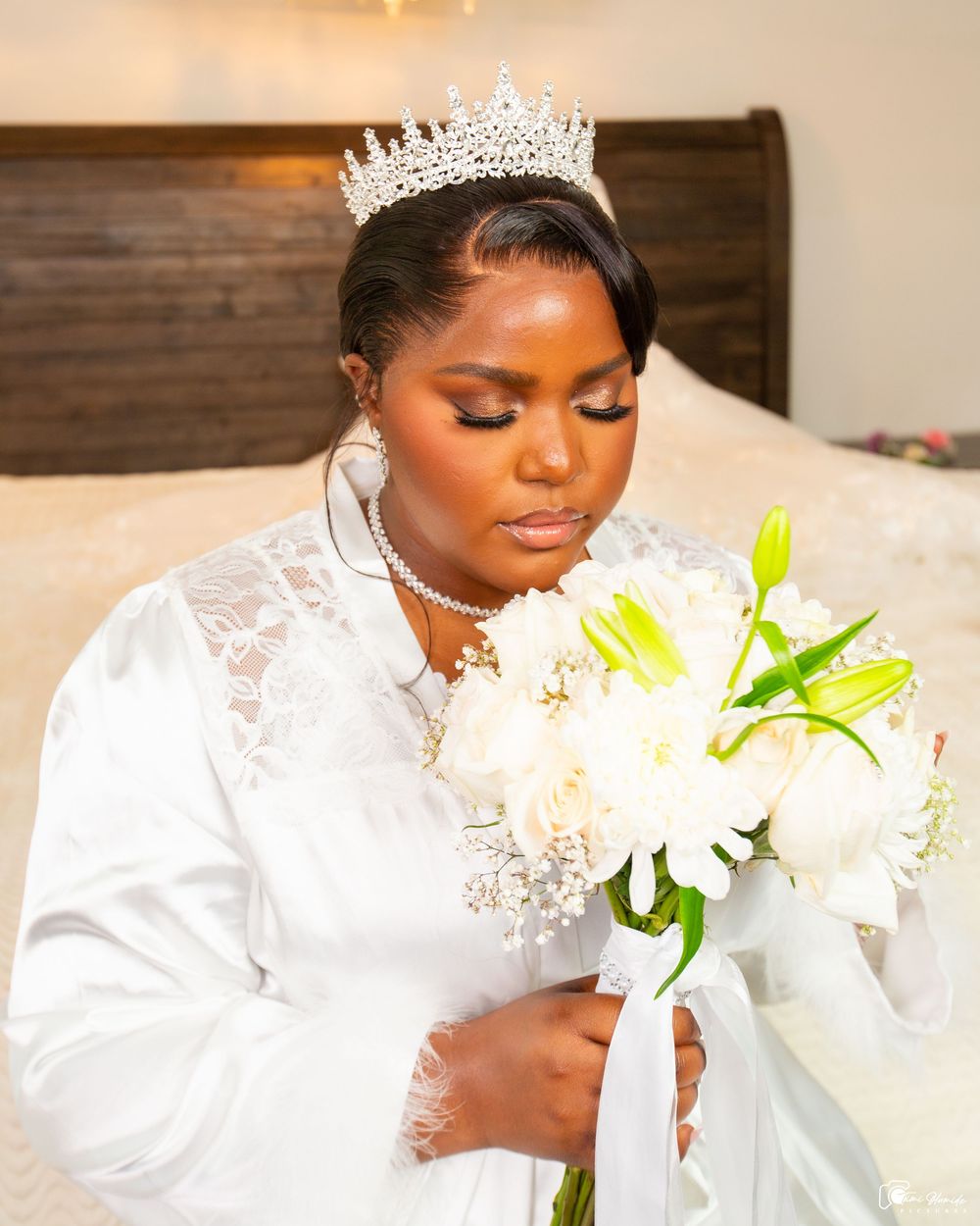 Elegant bride in white lace gown with crystal tiara poses with white and yellow lilies at an indoor photoshoot.