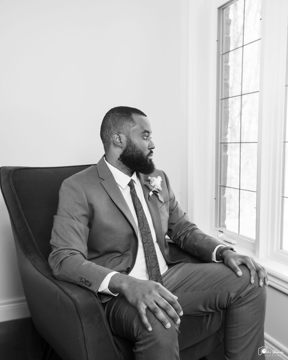 A contemplative man in a grey suit sits in a dark chair looking out a window in black and white photography.