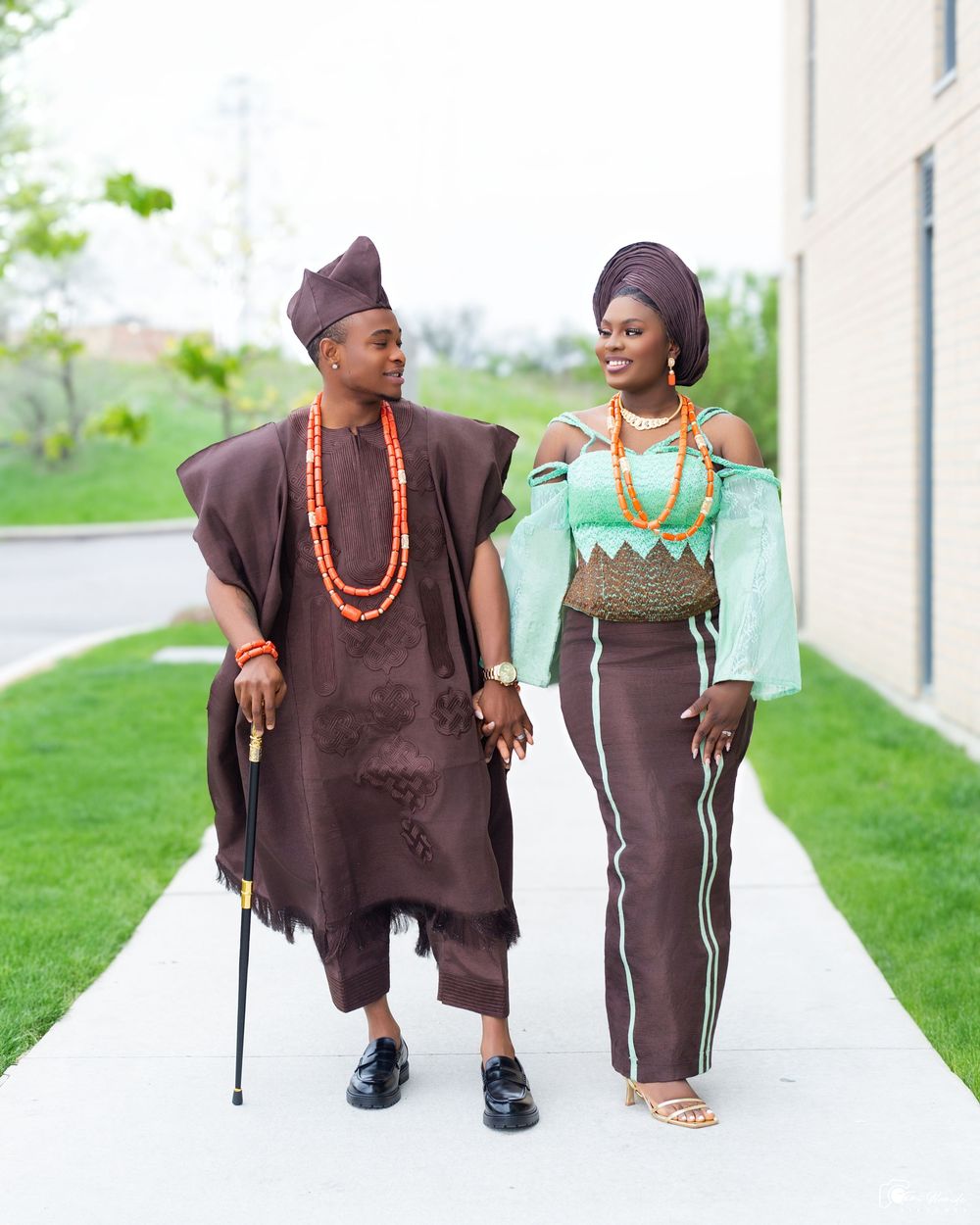 A couple wearing traditional brown Nigerian attire with beaded accessories pose for engagement photos by a white building.