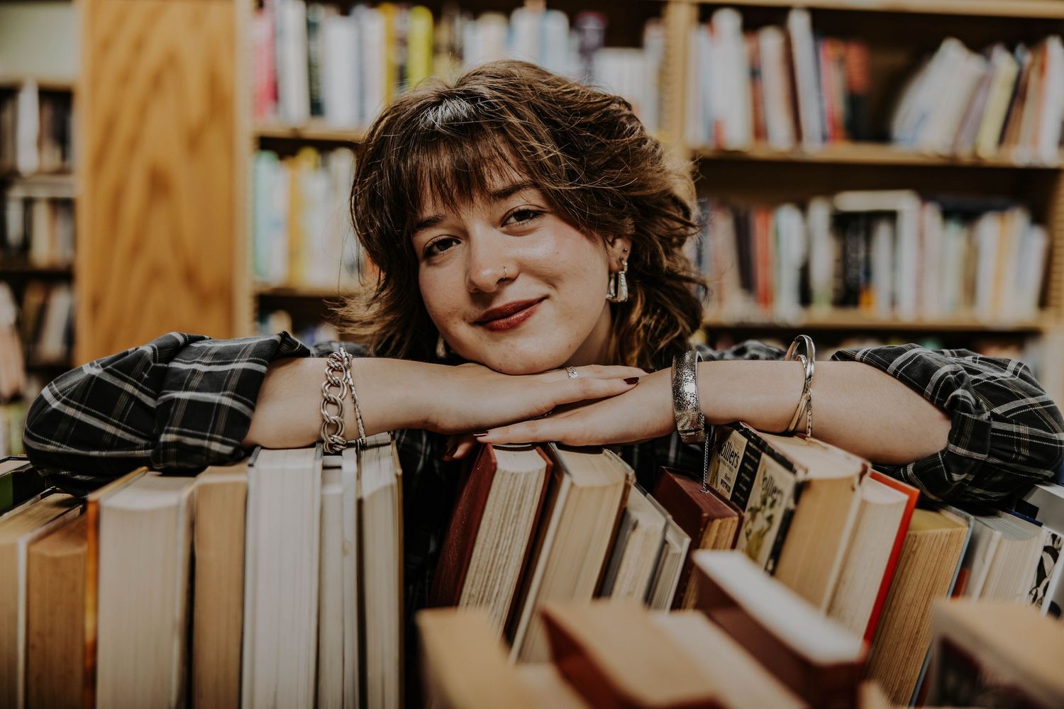 A smiling person rests their head on hands while leaning over stacks of books in a library.