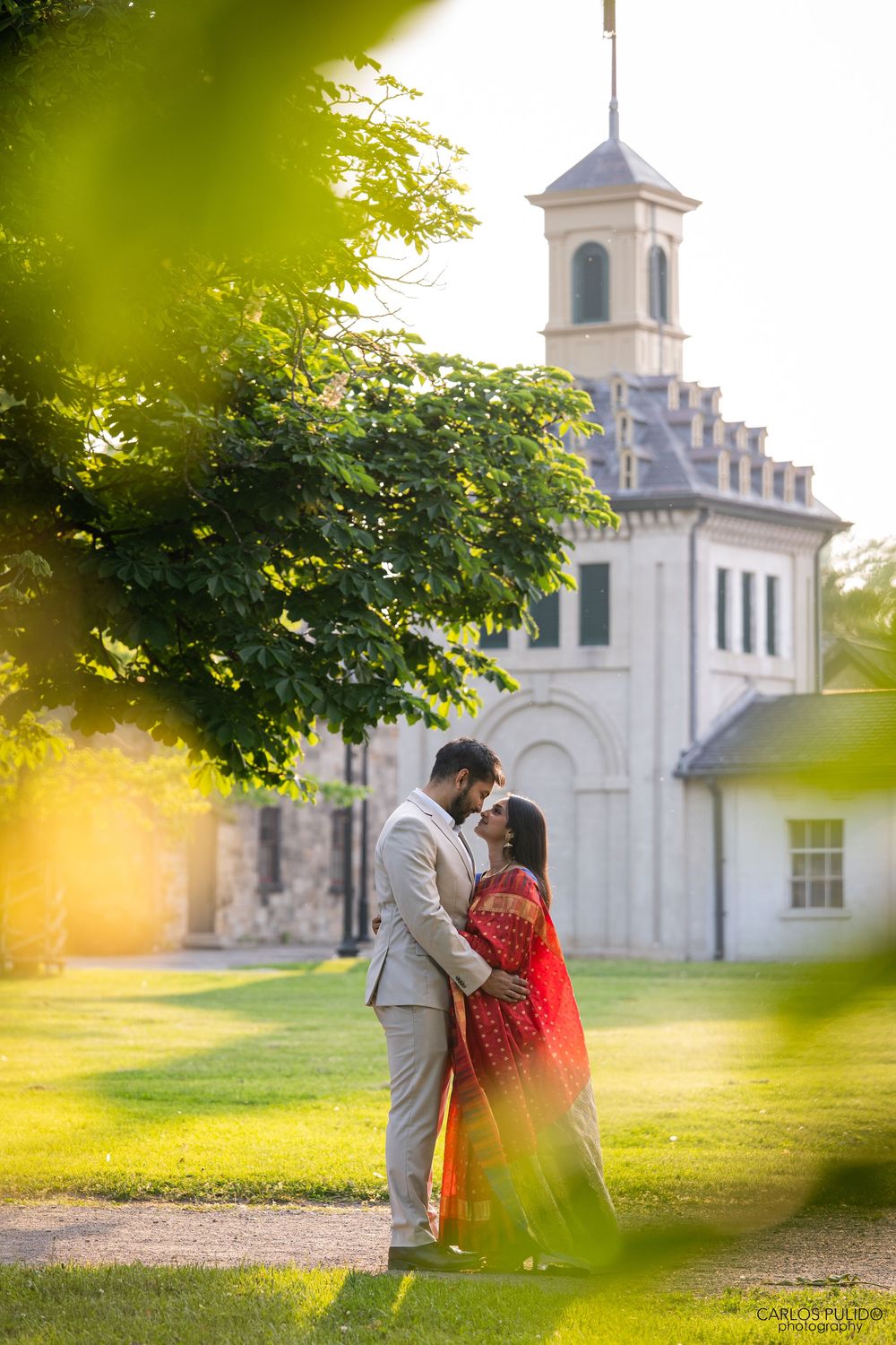 Couple shares romantic moments by a white church steeple at golden hour in a serene park setting.