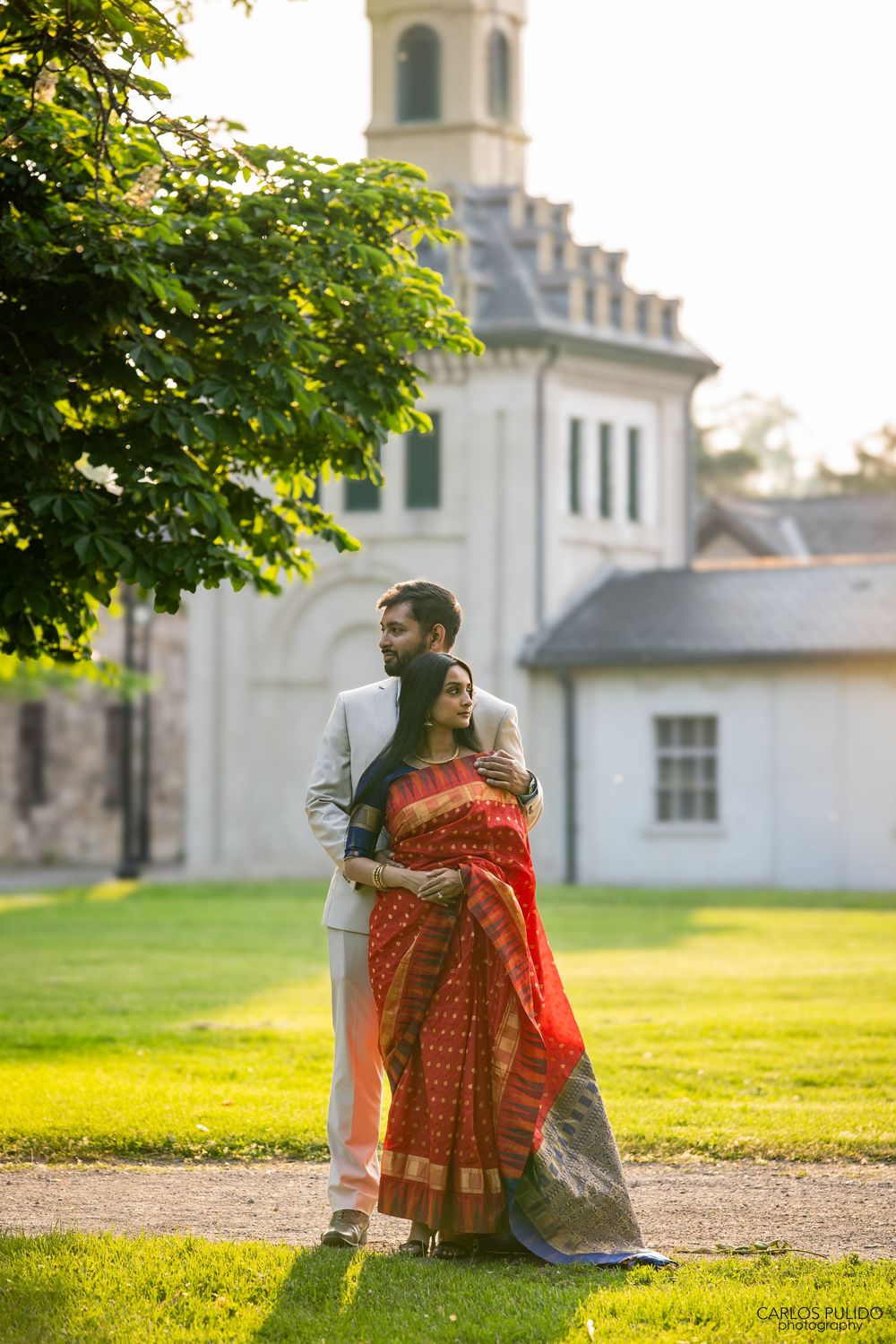 Couple in formal attire embracing outside historic church building with sunlit garden in golden evening light.
