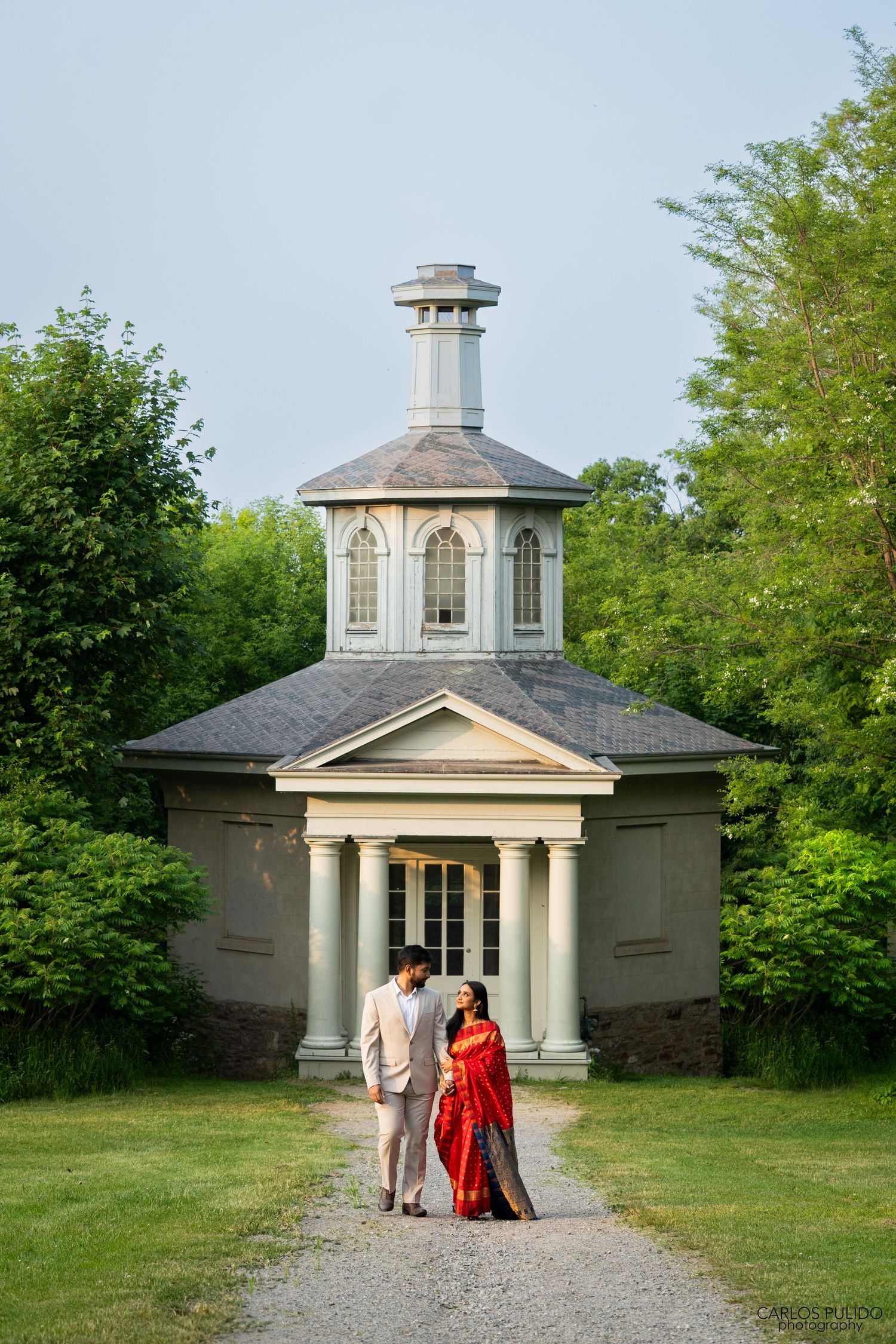 A couple walks along a gravel path toward an elegant white neoclassical chapel with columns and a cupola in a garden setting.