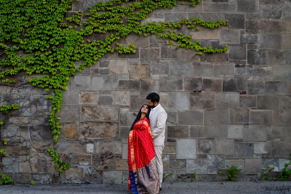 A couple embraces near an old stone wall with climbing ivy, the woman wearing a vibrant red dress.