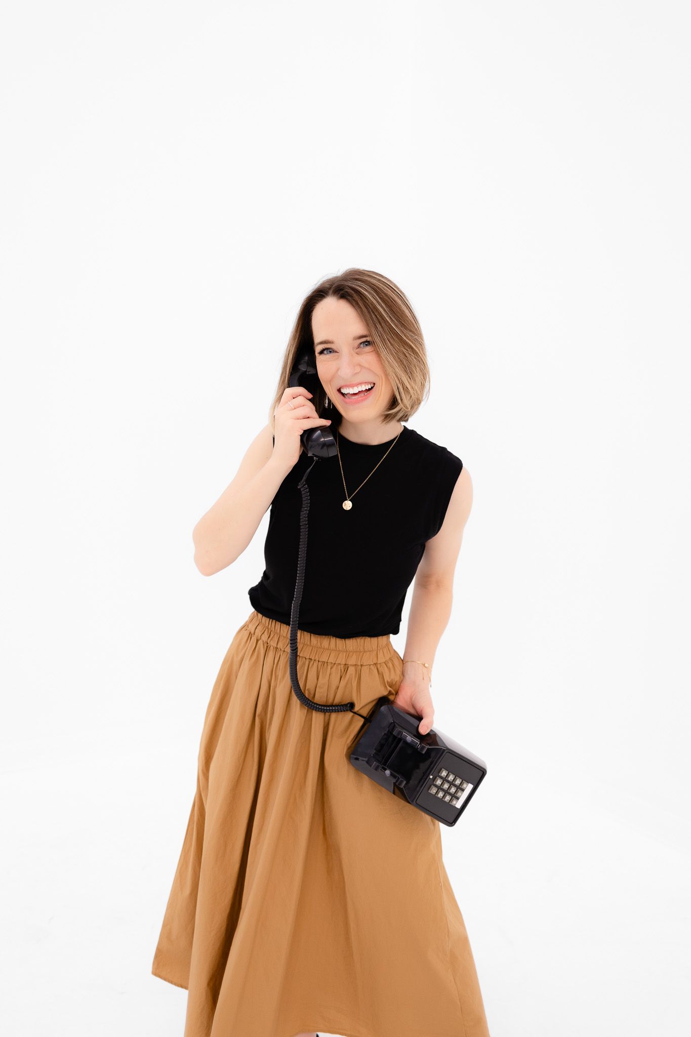 Person in a black sleeveless top and tan skirt holds a vintage telephone while smiling against white background.