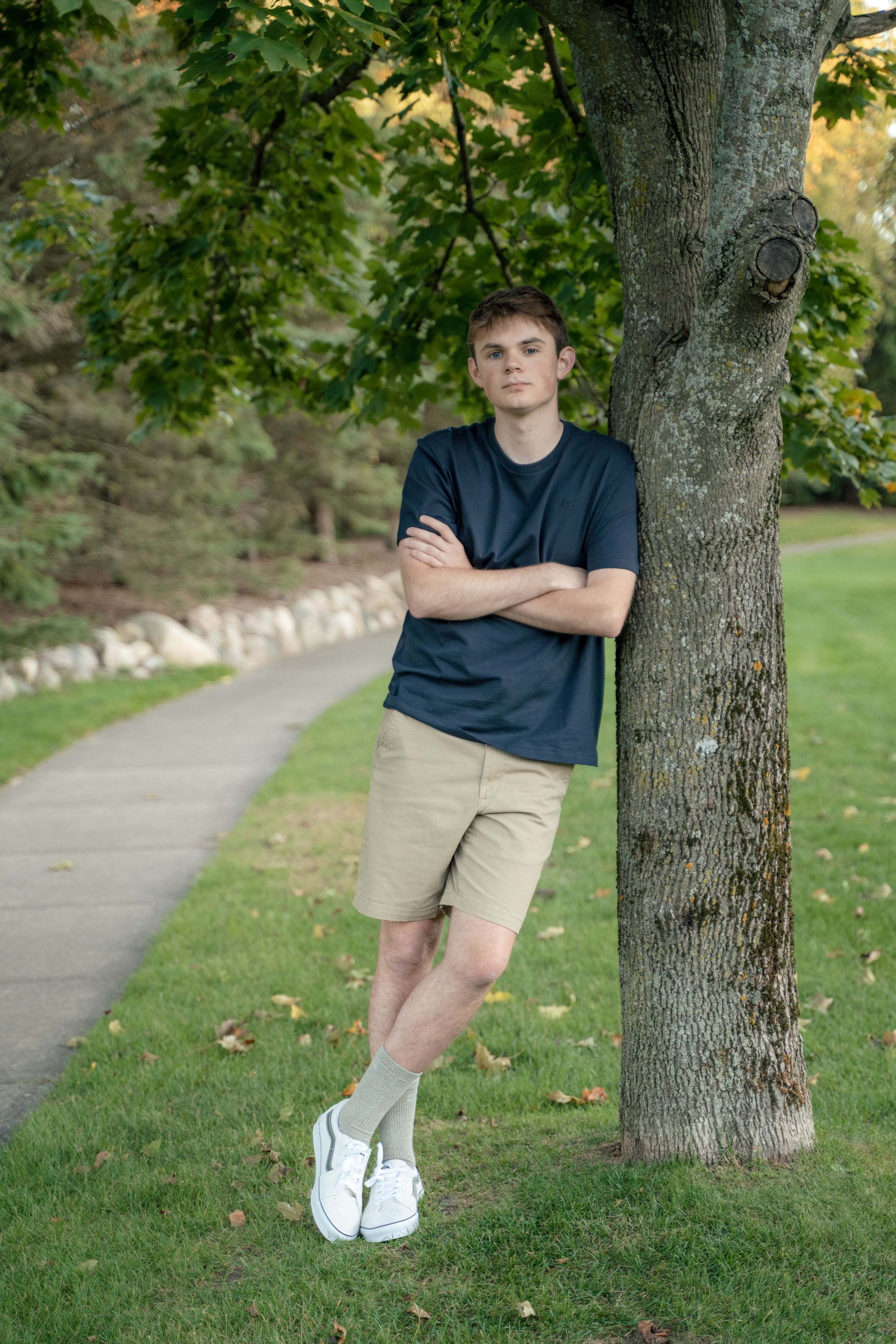 A person in a navy shirt and khaki shorts leans against a tree trunk along a paved path on a grassy lawn.