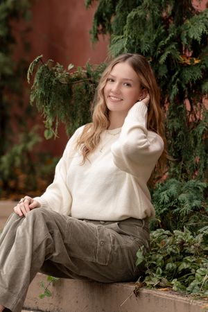A person wearing a white sweater and cargo pants poses outdoors near evergreen foliage against a warm-toned background.