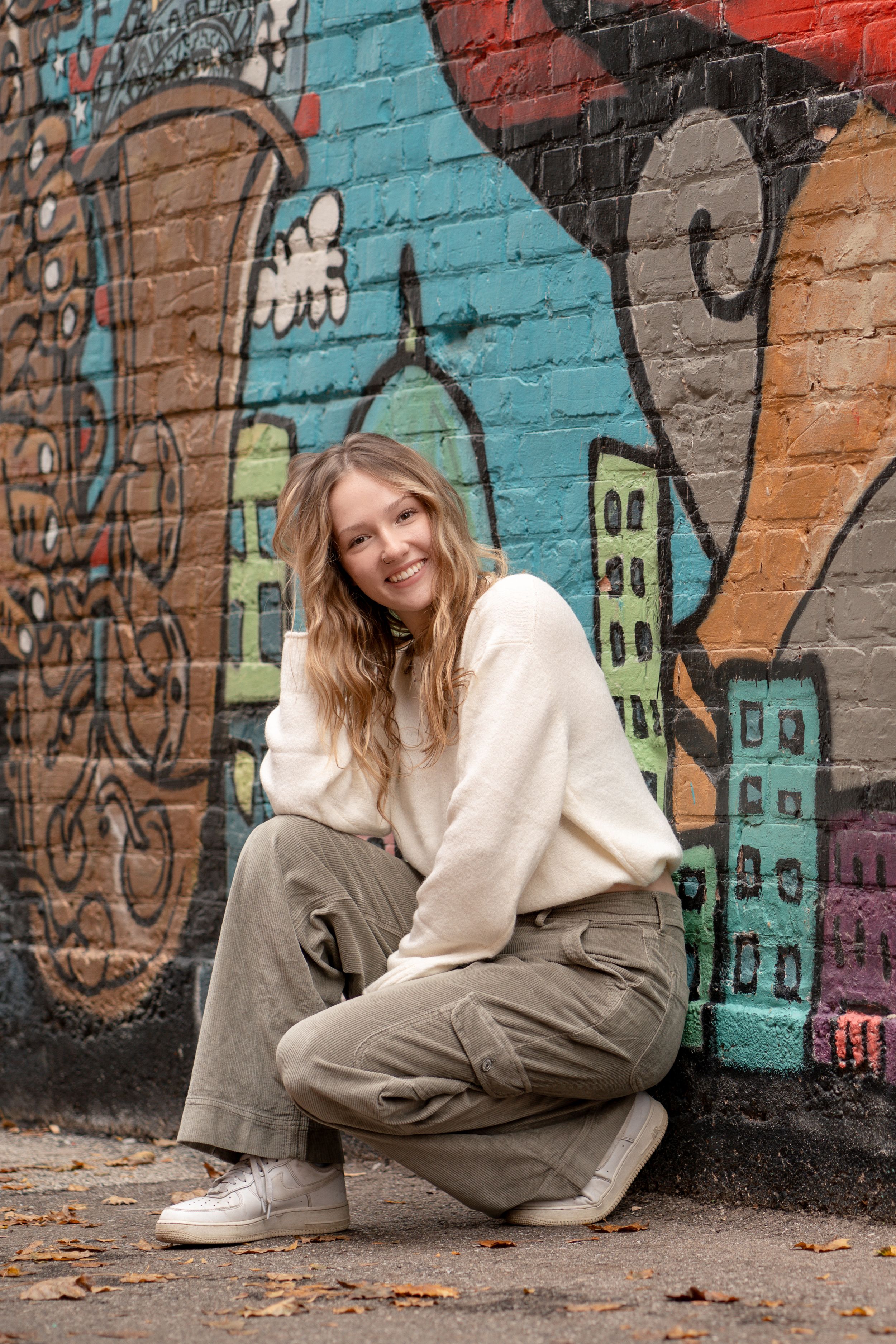 Young model in cream sweater and grey pants poses against colorful graffiti wall during casual urban photoshoot.