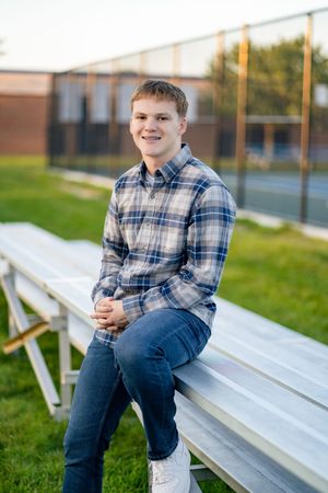 Someone in a plaid flannel shirt and jeans sits casually on aluminum bleachers at an outdoor sports field.
