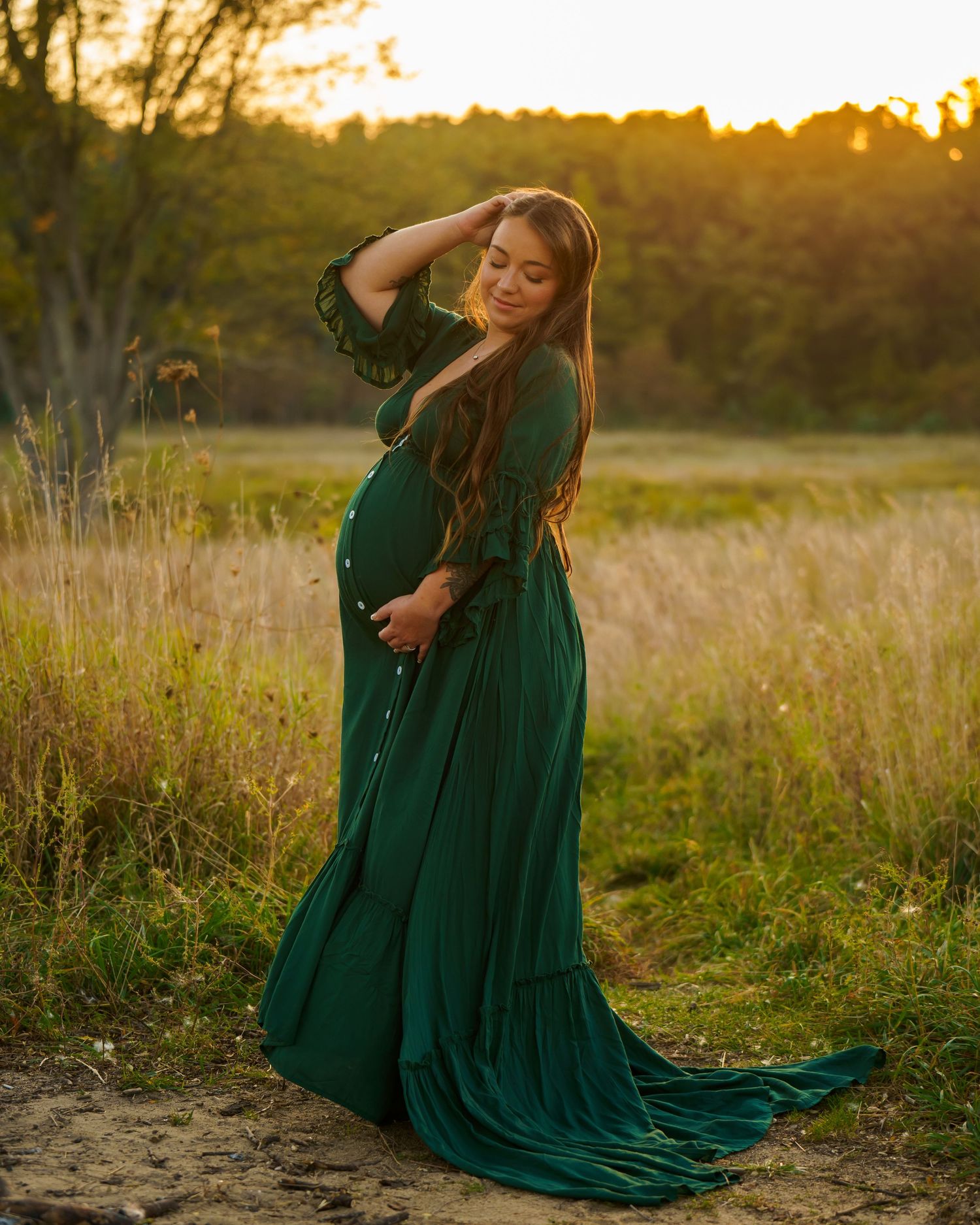 Expectant mother in flowing emerald green dress poses gracefully in a sunlit meadow at golden hour.