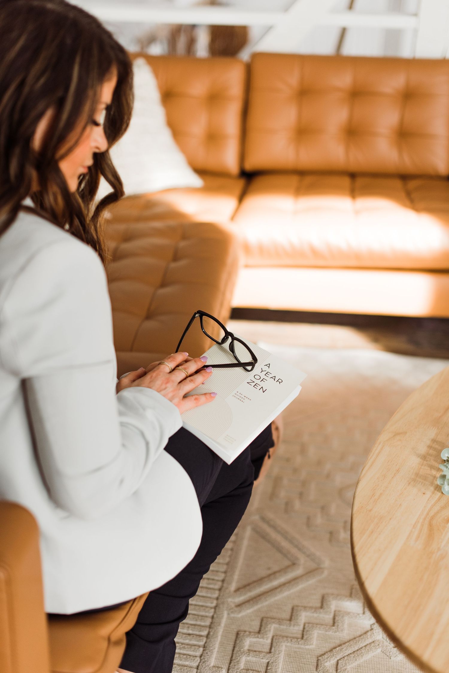 A person wearing a white blazer sits on a brown leather couch while writing in a notebook.