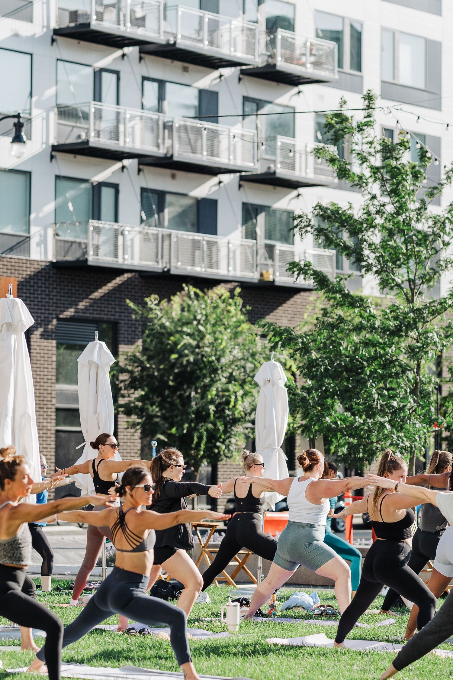 Group of fitness enthusiasts practicing yoga warrior poses in an outdoor class with modern apartment building backdrop.
