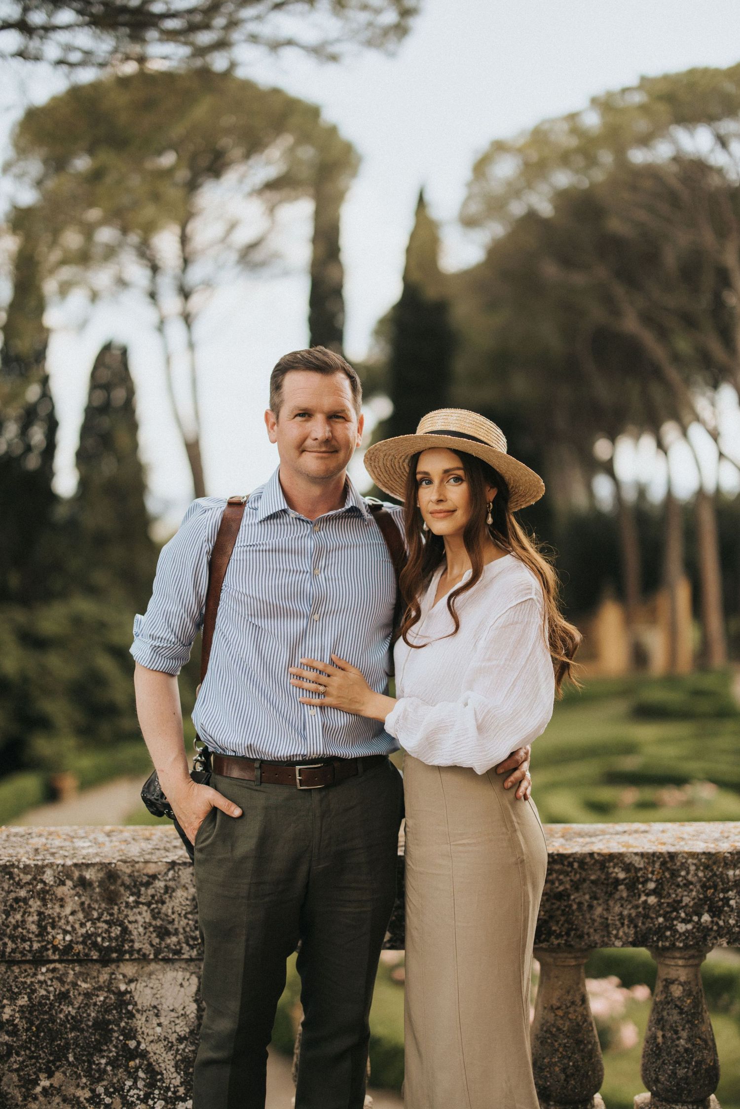 A couple in elegant attire poses together on a stone balcony with Italian cypress trees in the background.