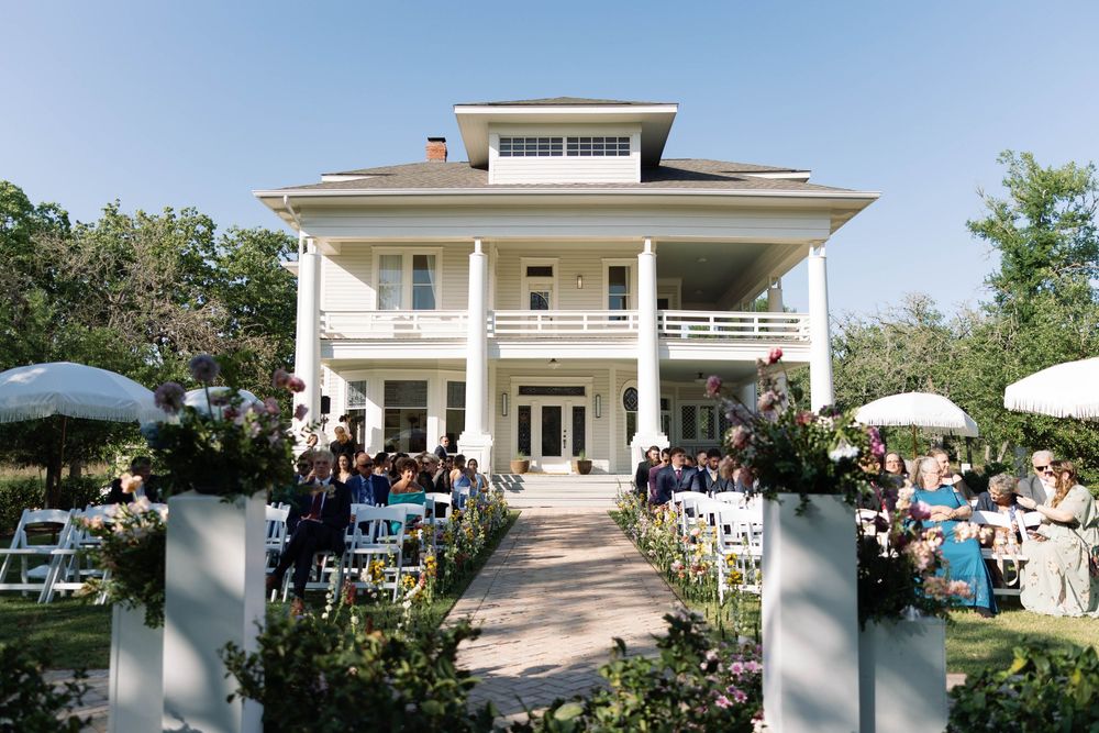 A grand white two-story mansion with classic columns hosts an outdoor wedding ceremony with guests seated in rows on the lawn.