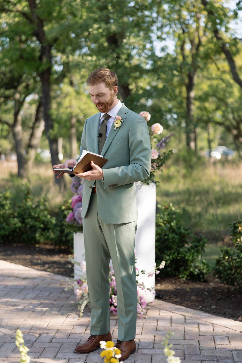 A person in a mint green suit reads from a book while standing on a paved garden path surrounded by flowers and trees.