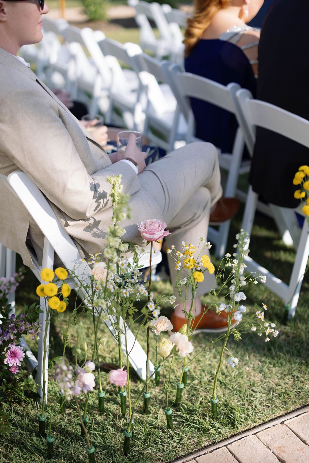 Wildflowers and pink roses line the aisle at an outdoor wedding ceremony with white chairs on grass.