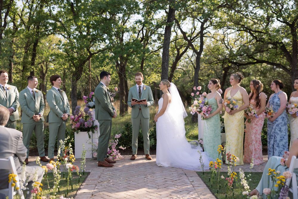 Wedding ceremony guests gather outdoors under lush green trees during a sunny summer celebration.