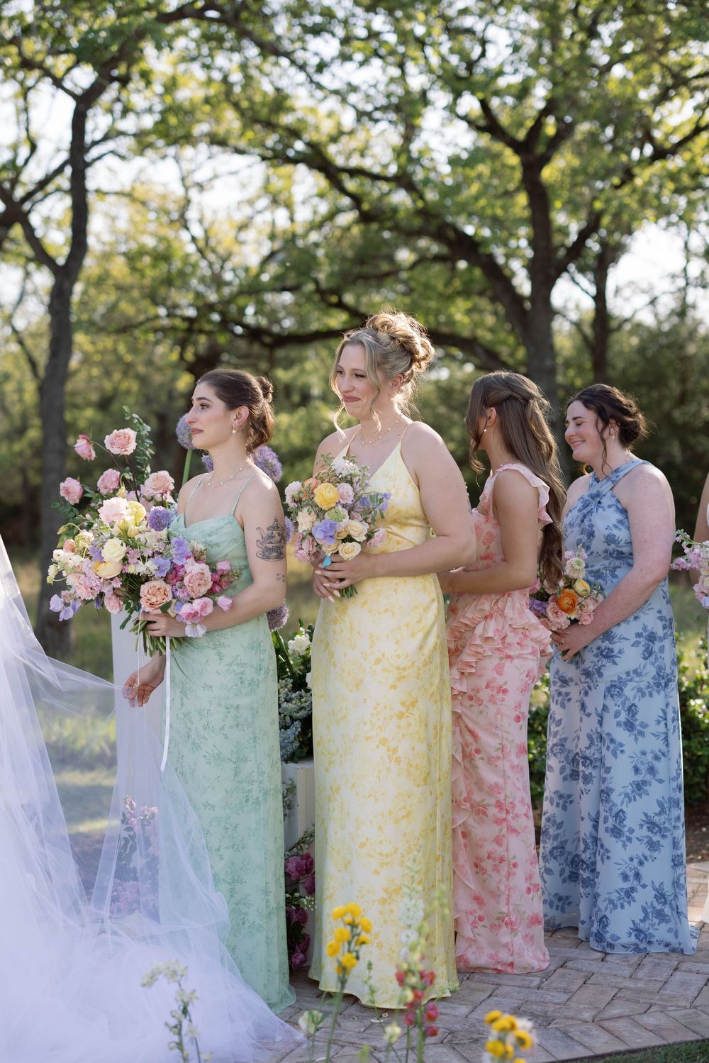 Bridesmaids in pastel floral dresses stand together holding colorful bouquets during an outdoor garden wedding ceremony.