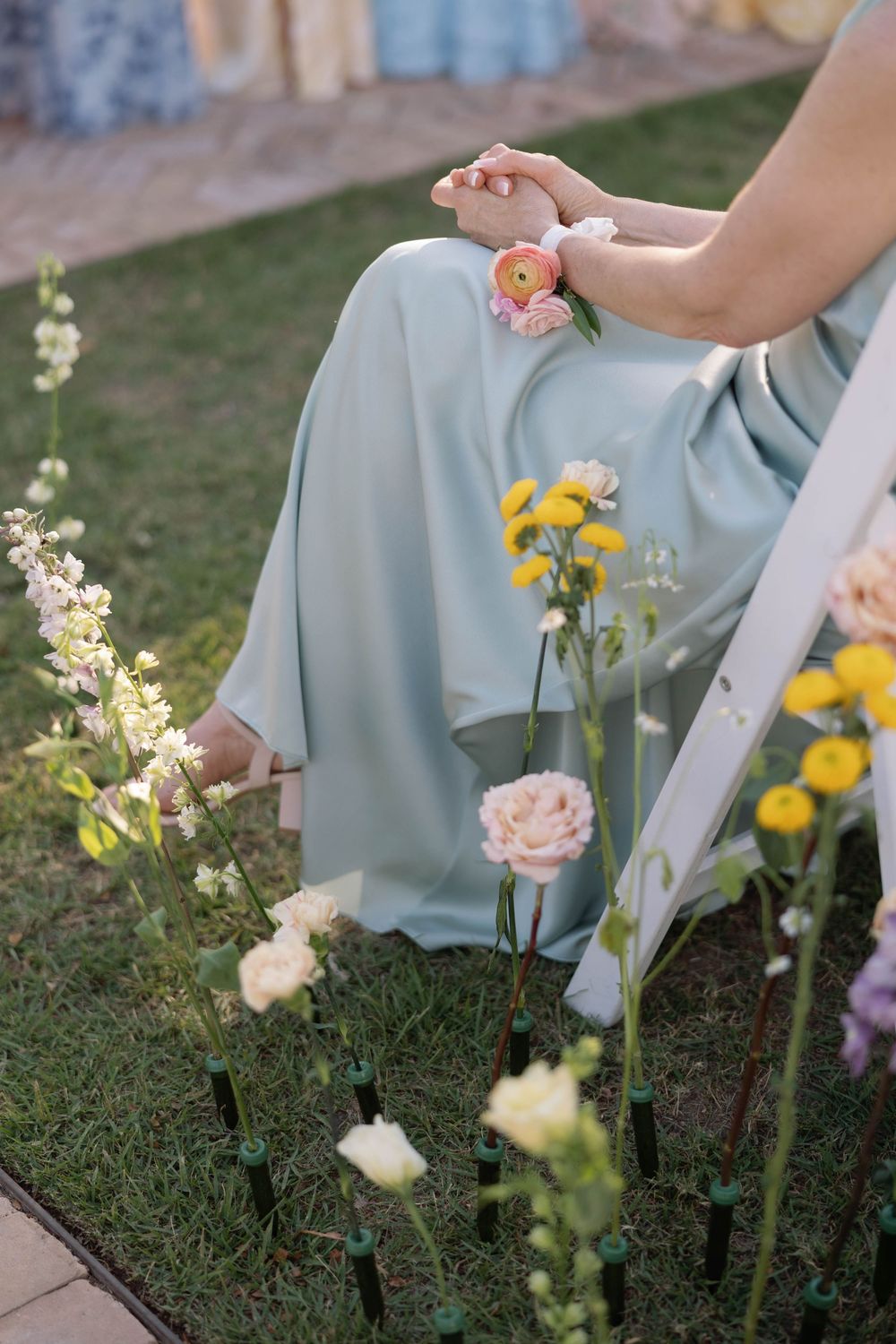 Person in a flowy mint green dress sits among blooming garden flowers including yellow and pink blossoms in soft lighting.