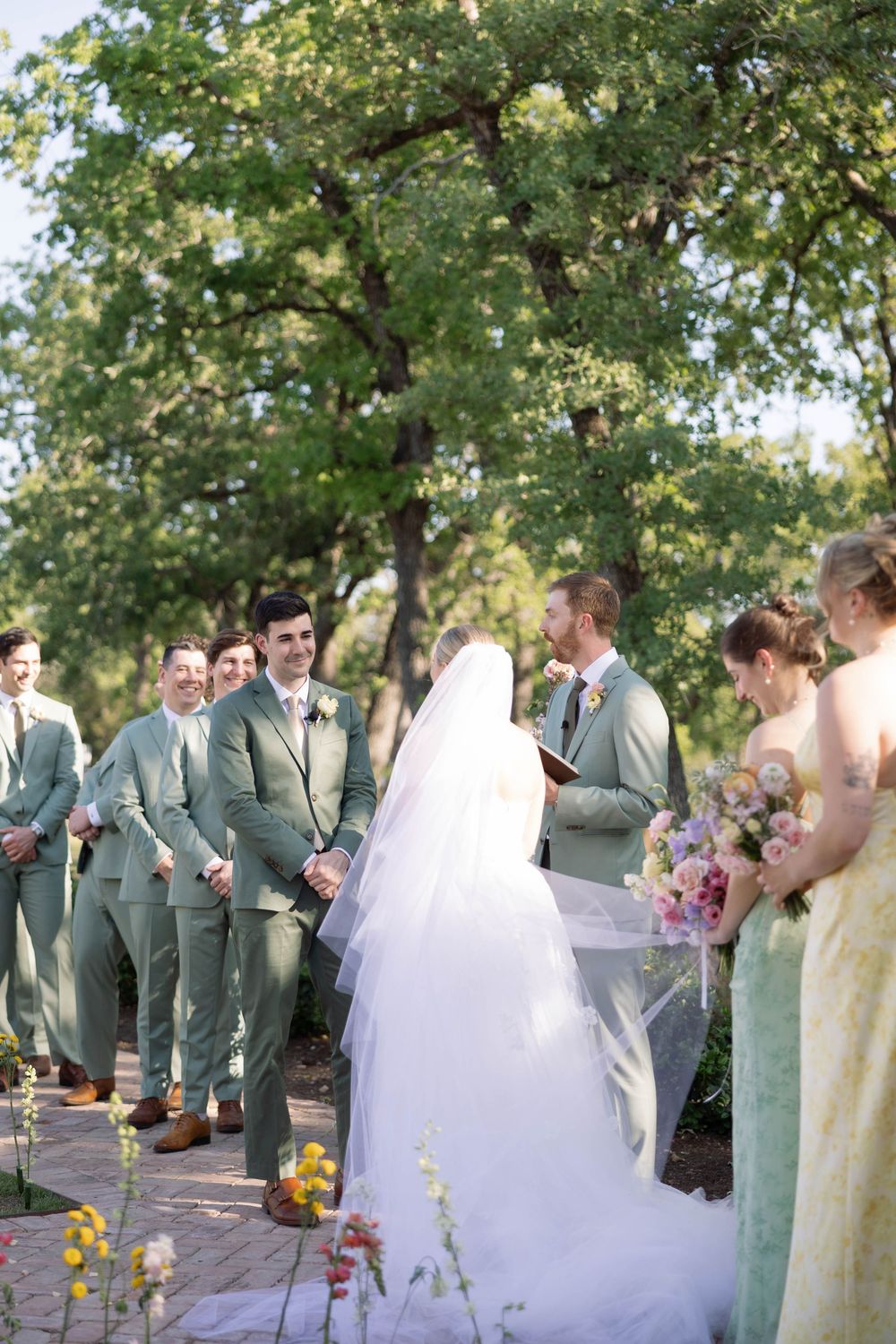 Wedding party in grey suits and mint green dresses gather outdoors under trees for cheerful group photos.