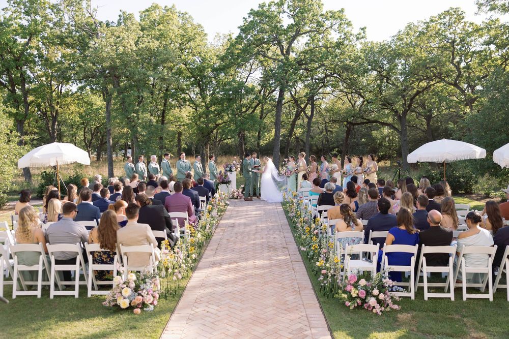 An outdoor wedding ceremony setup with white chairs, umbrellas and a brick pathway aisle surrounded by green trees.