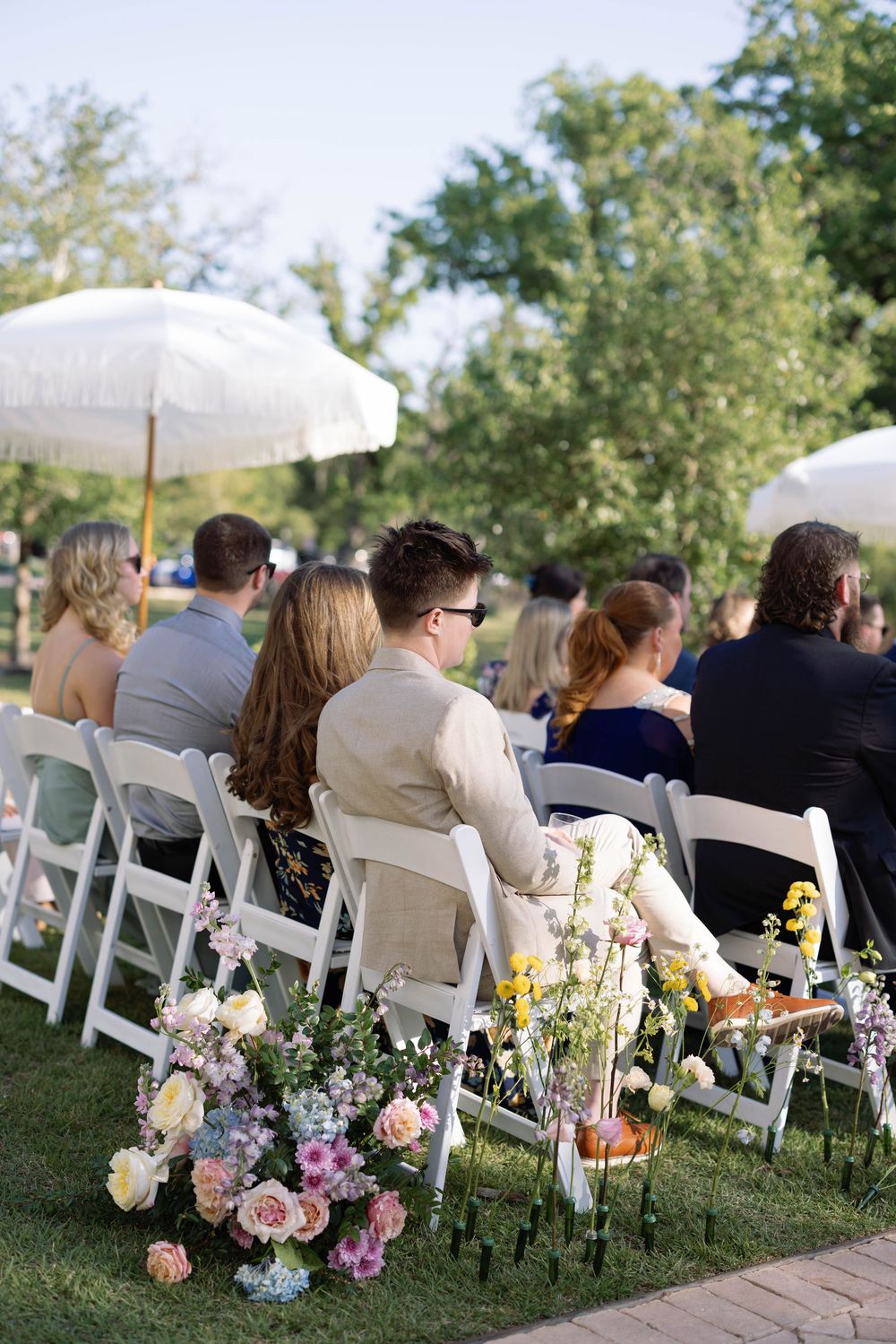 White chairs and floral arrangements line an outdoor garden ceremony space under white umbrellas on a sunny day.