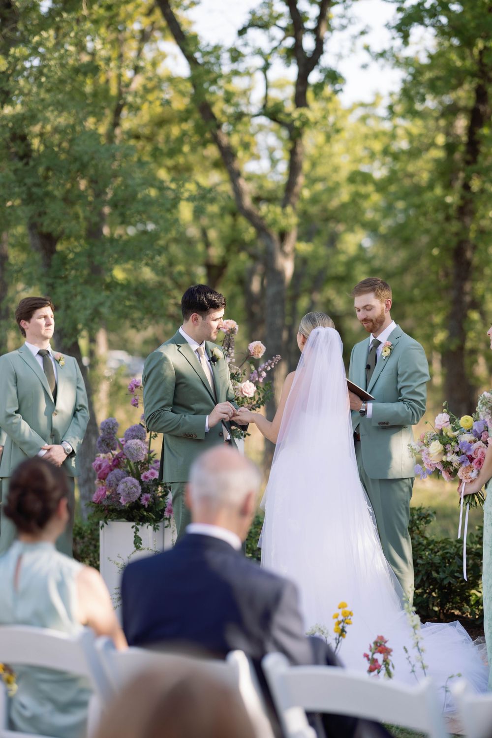 An outdoor wedding ceremony takes place in a wooded setting with sage green suits and colorful floral arrangements.