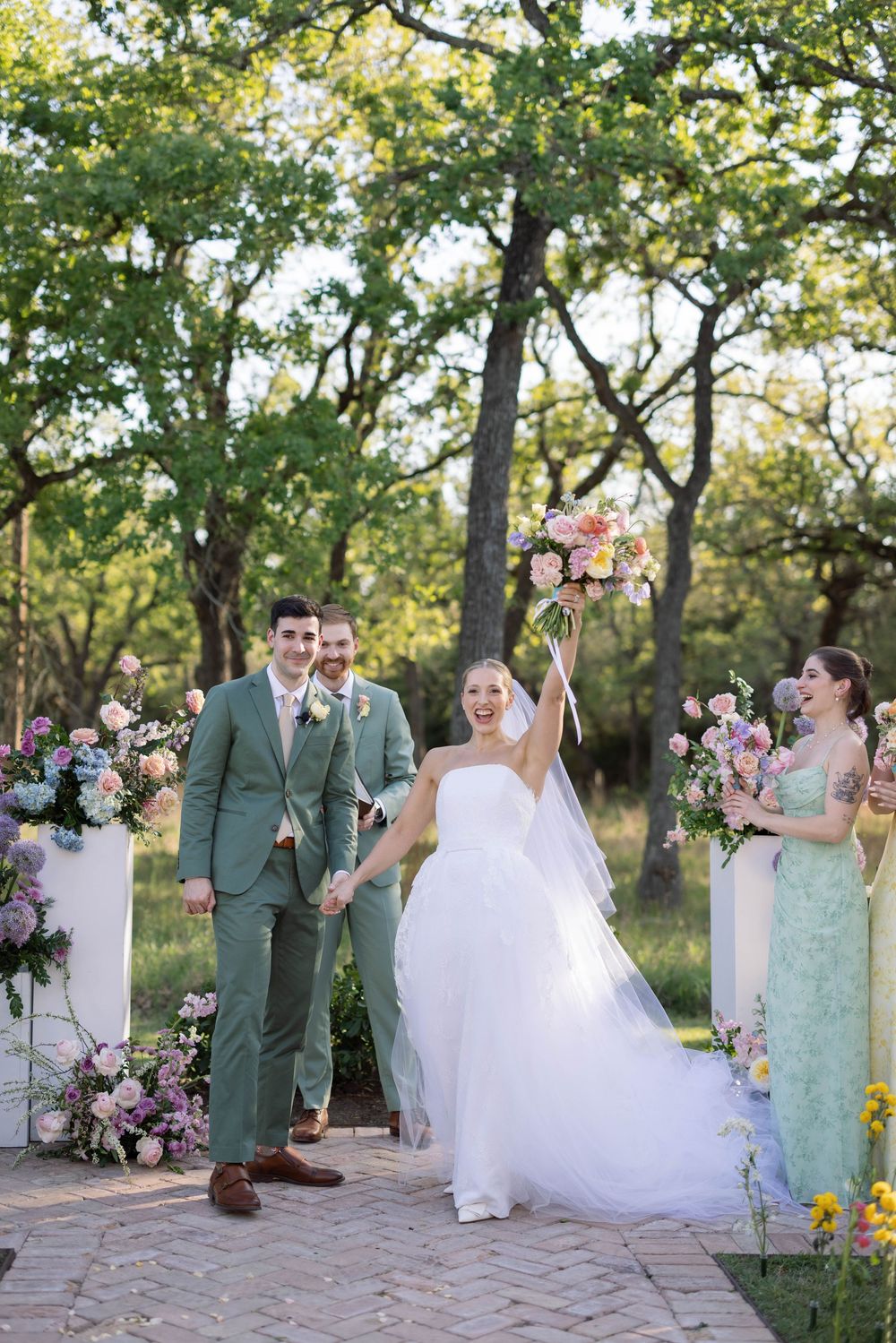 Outdoor wedding ceremony on brick path with floral arch decorations and pink blooms in a wooded garden setting.