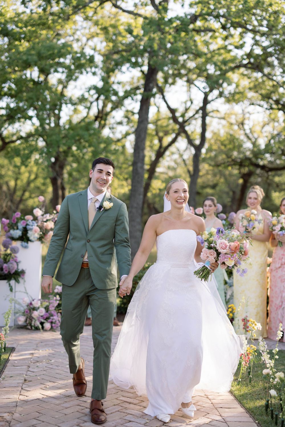 A couple walks down a sunlit garden path after their wedding ceremony while attendants follow behind with bouquets.