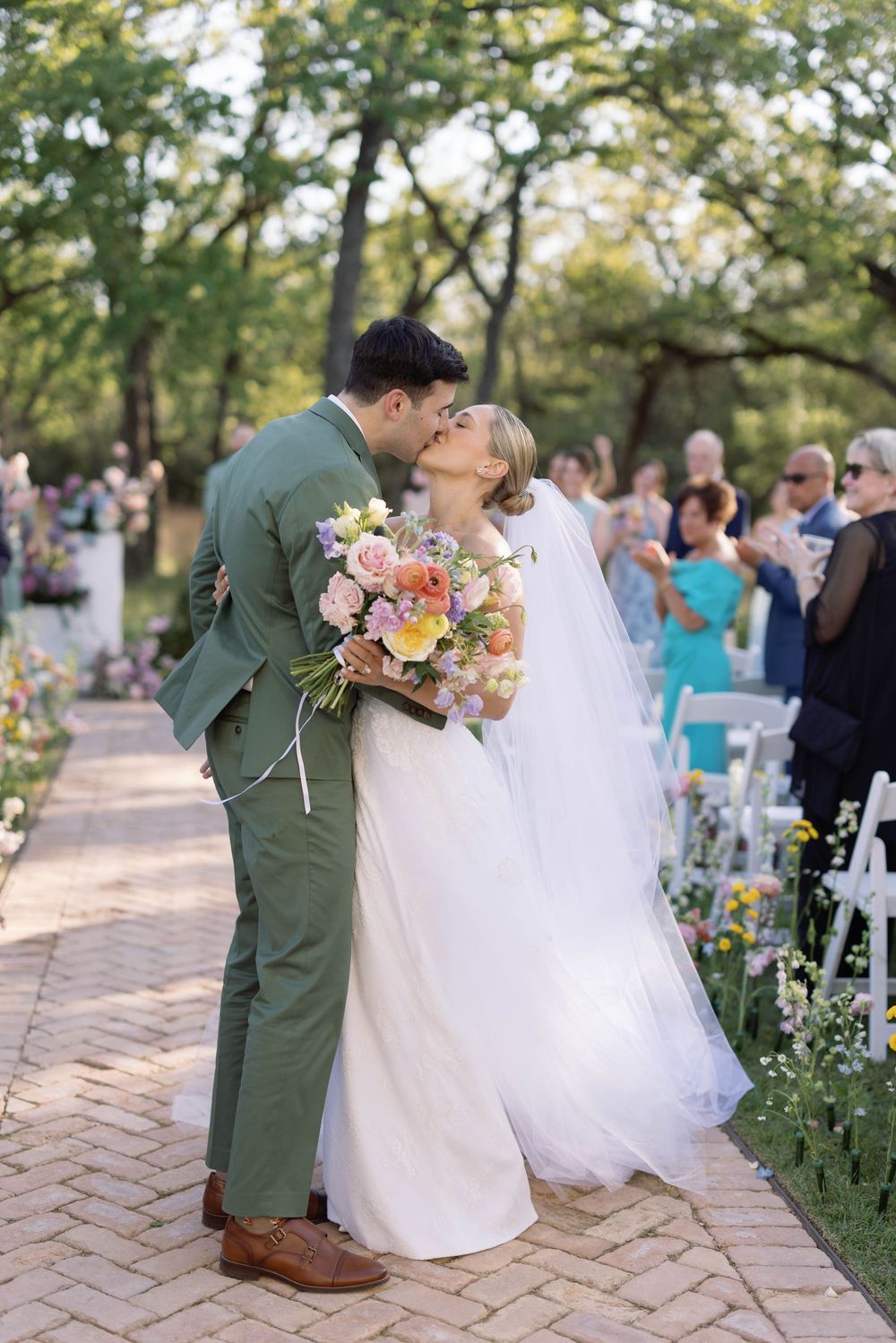 A bride and groom share a romantic kiss on a brick pathway surrounded by wedding guests and floral arrangements.