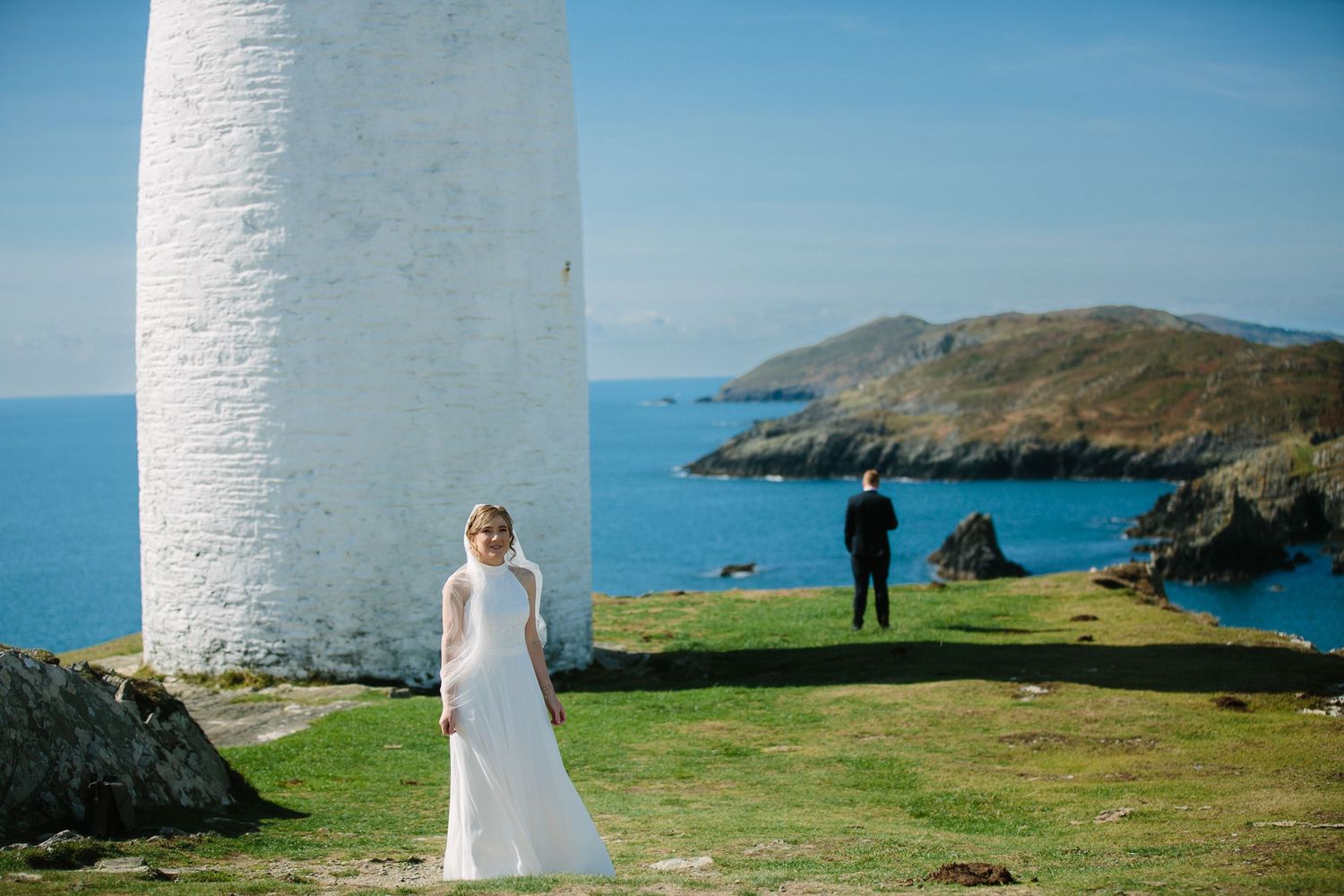 A person stands in a white dress near a lighthouse with coastal cliffs and ocean views in Ireland.