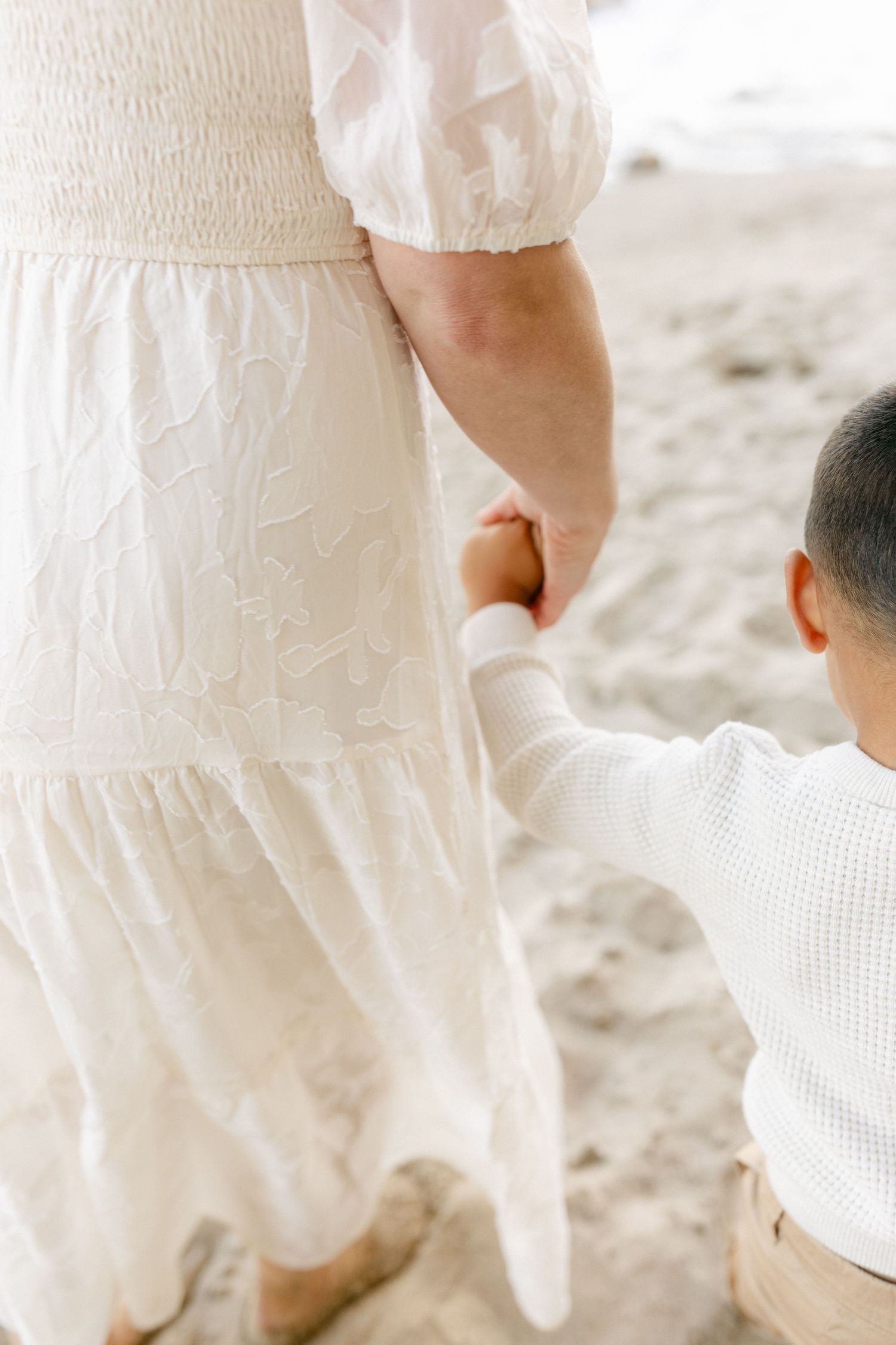 A close-up view of a child in white clothing reaching for someone in a flowing white dress on a sandy beach.