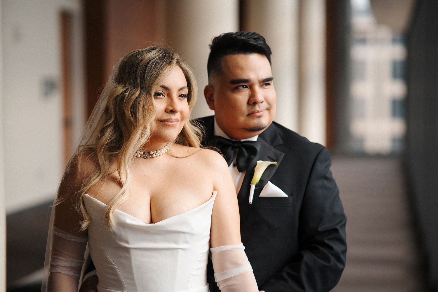 A couple in formal wedding attire: a sleek white gown with sheer sleeves and a classic black tuxedo.