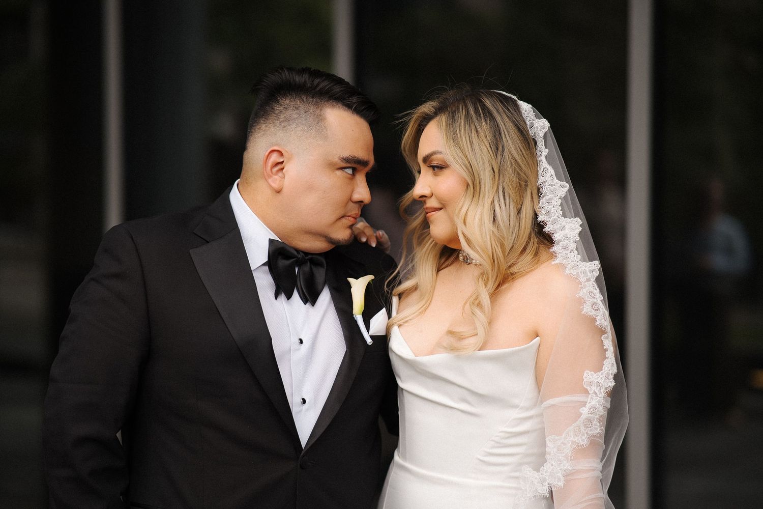 A romantic couple shares an intimate moment in elegant wedding attire with a black tuxedo and white gown with lace veil.