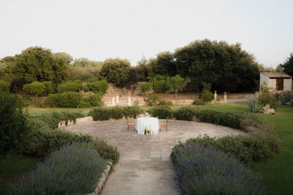 Stone pathway lined with lavender leads to a tranquil garden seating area surrounded by lush greenery and trees.