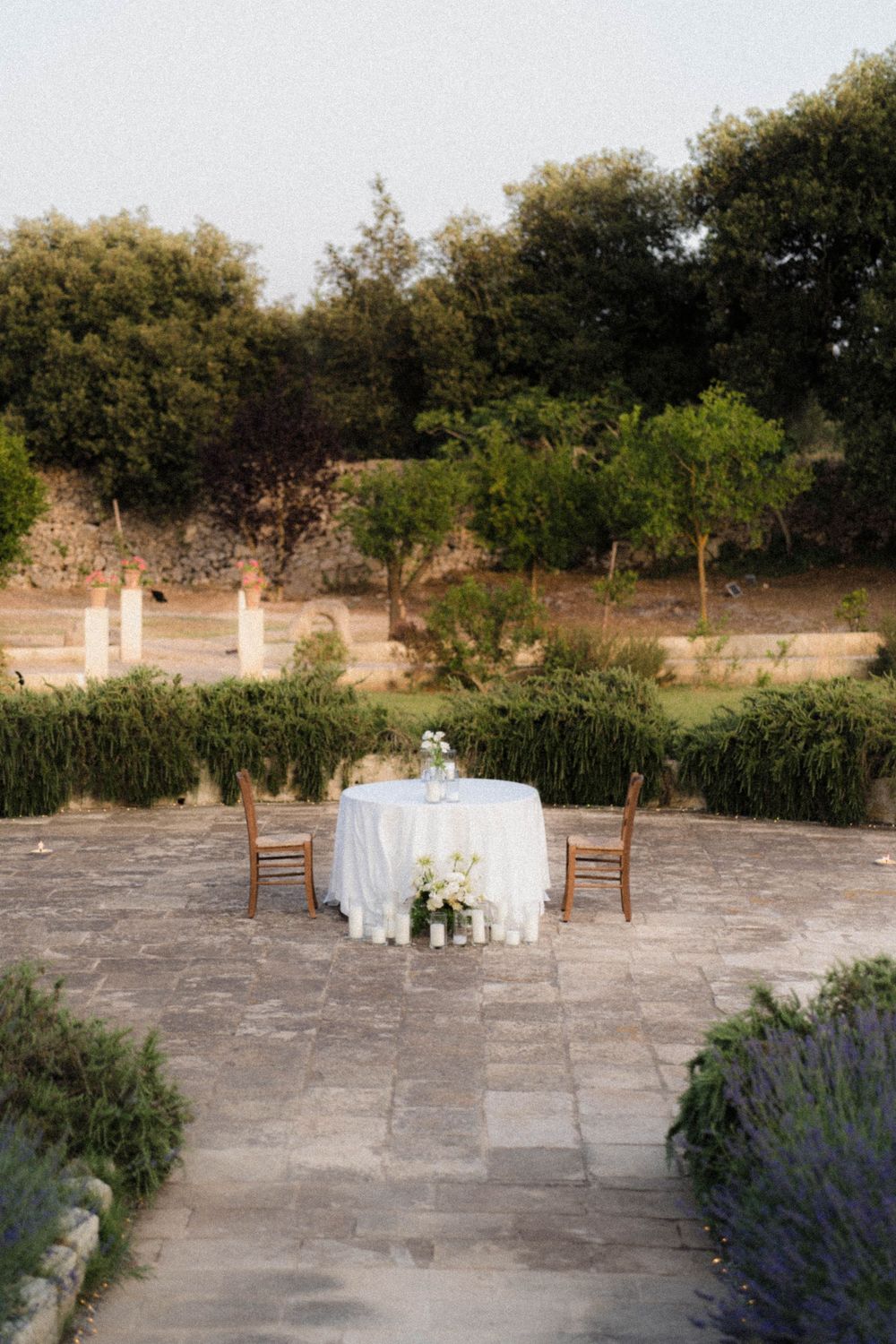 Intimate garden setting with round table draped in white linen and wooden chairs surrounded by stone patio and lavender.