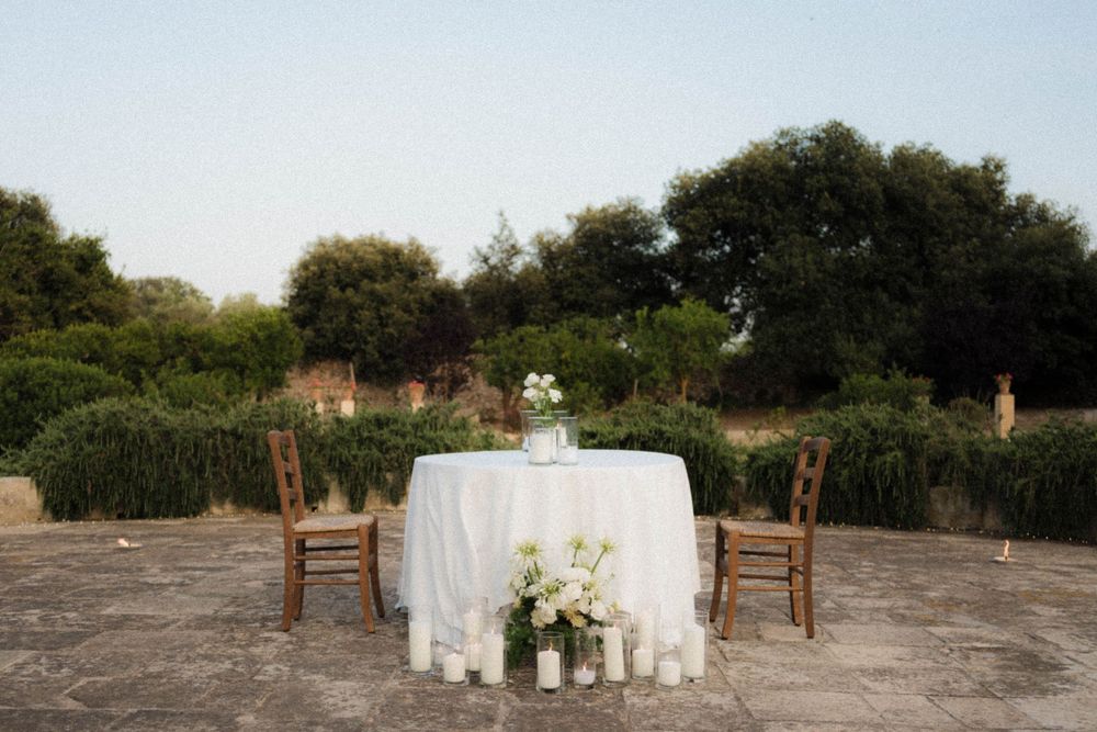A rustic outdoor wedding table setup with white linens and wooden chairs against a scenic natural backdrop at sunset.