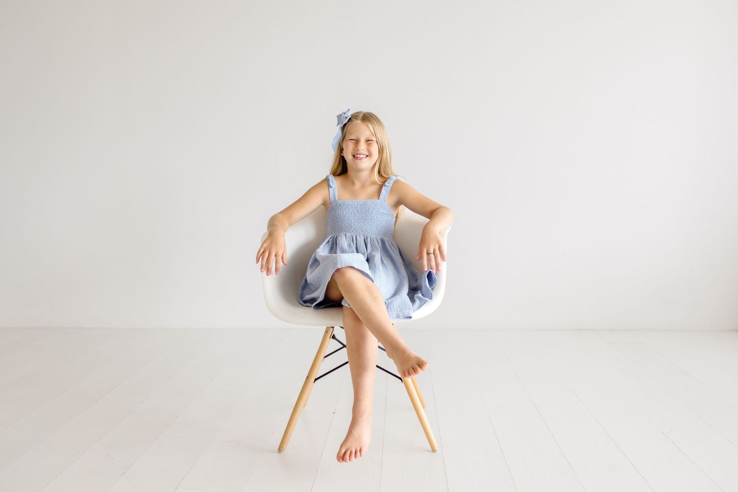 A person in a blue dress sits playfully on a modern white chair against a bright backdrop.