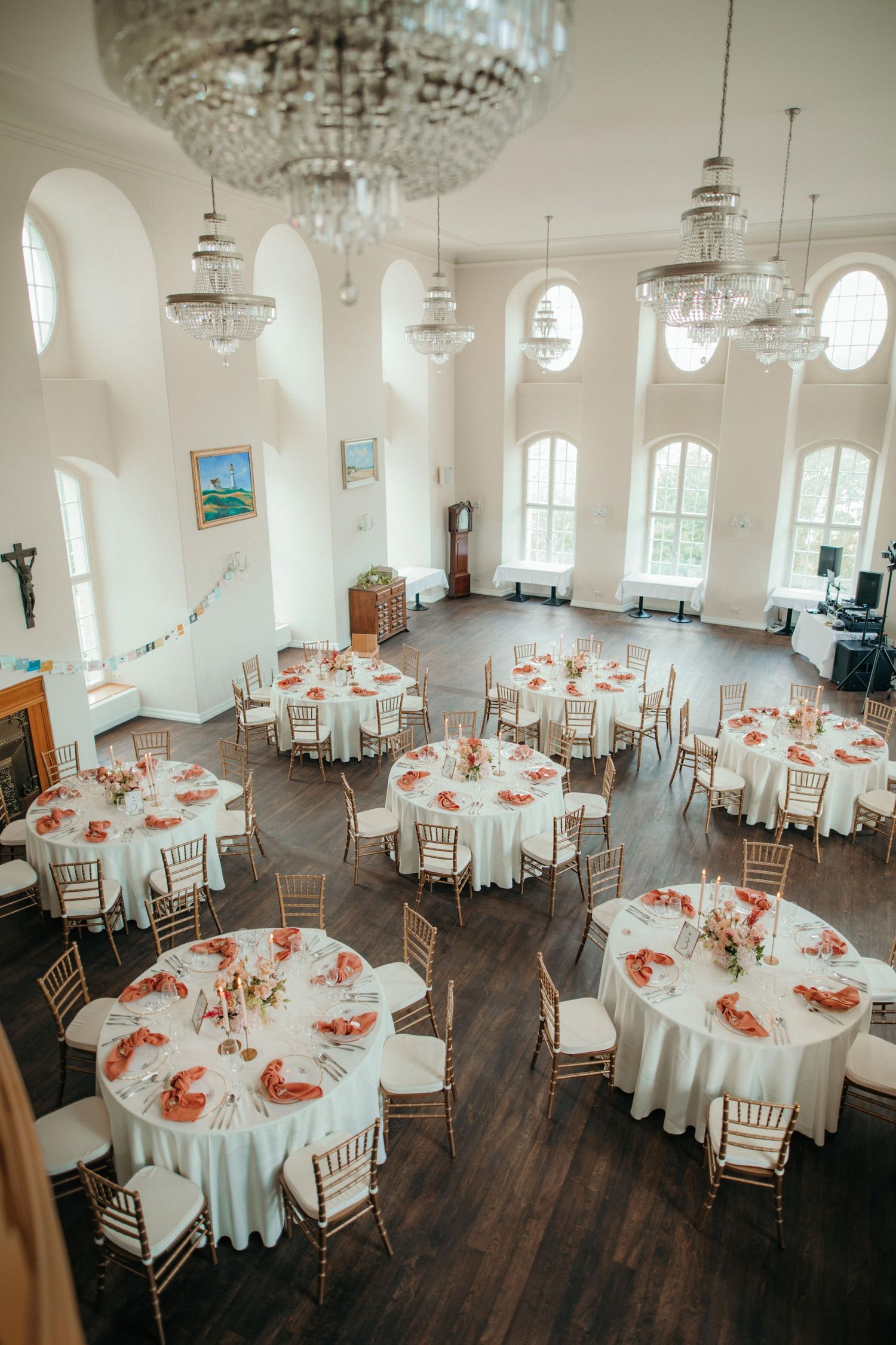 Elegant wedding reception venue with round tables decorated in white linens and coral centerpieces under crystal chandeliers.