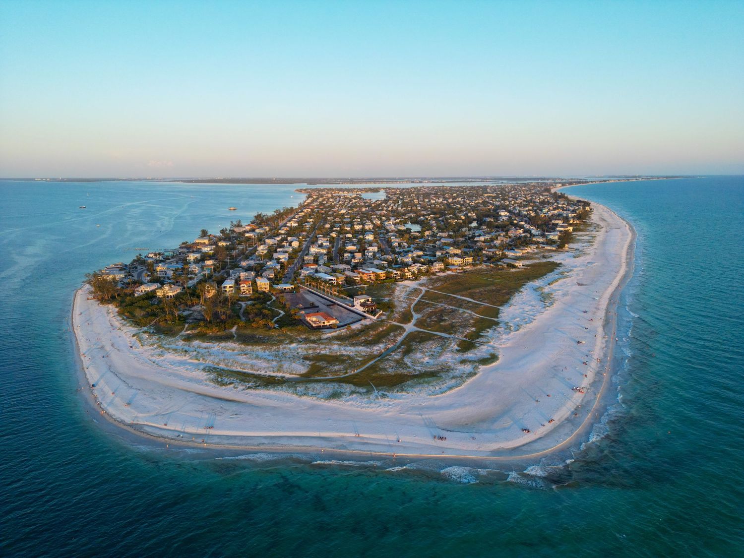 Aerial view of a curved beach peninsula with residential houses and turquoise waters surrounding the coastal community.
